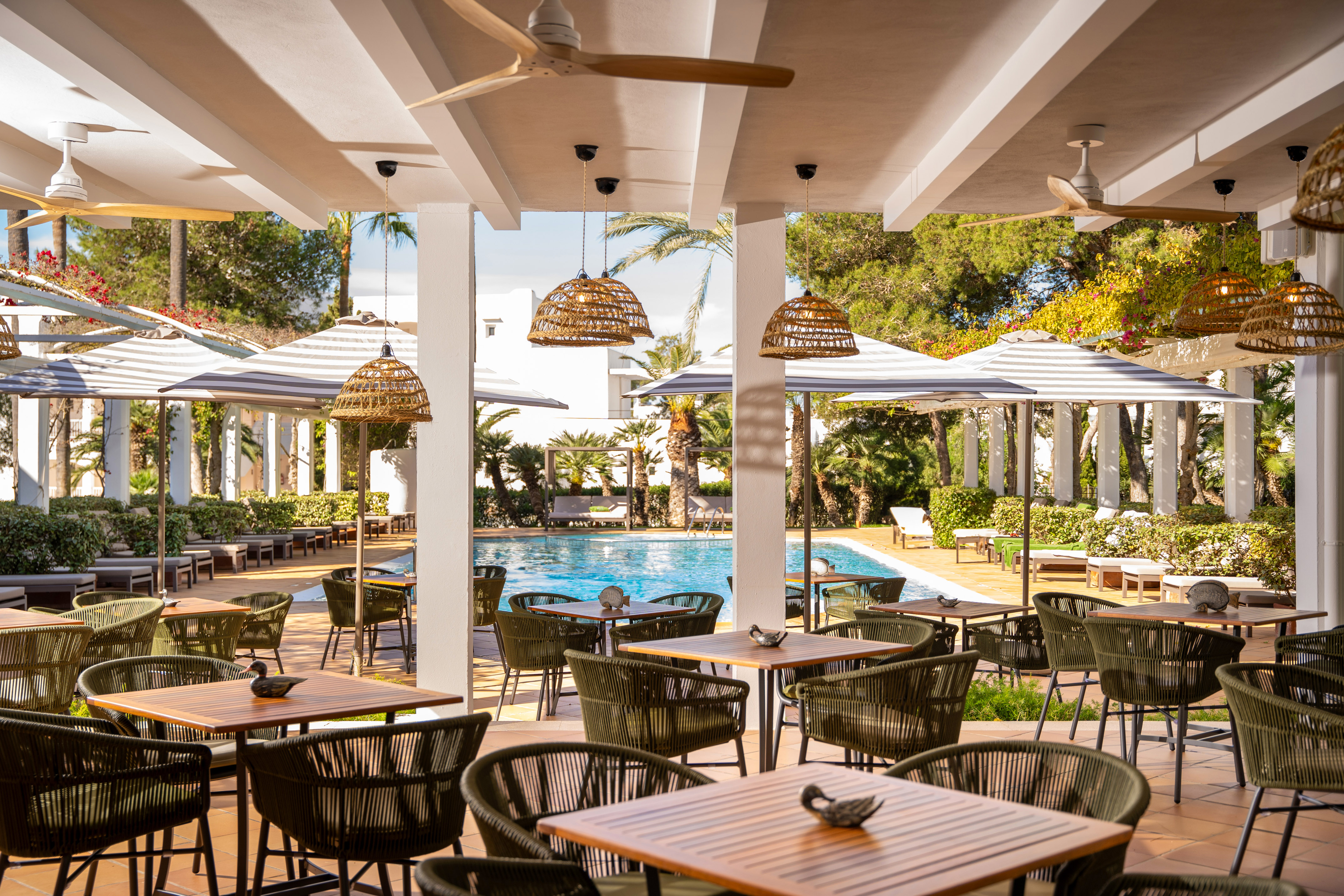 Poolside resort restaurant patio with woven chairs, wooden tables, and palm trees.