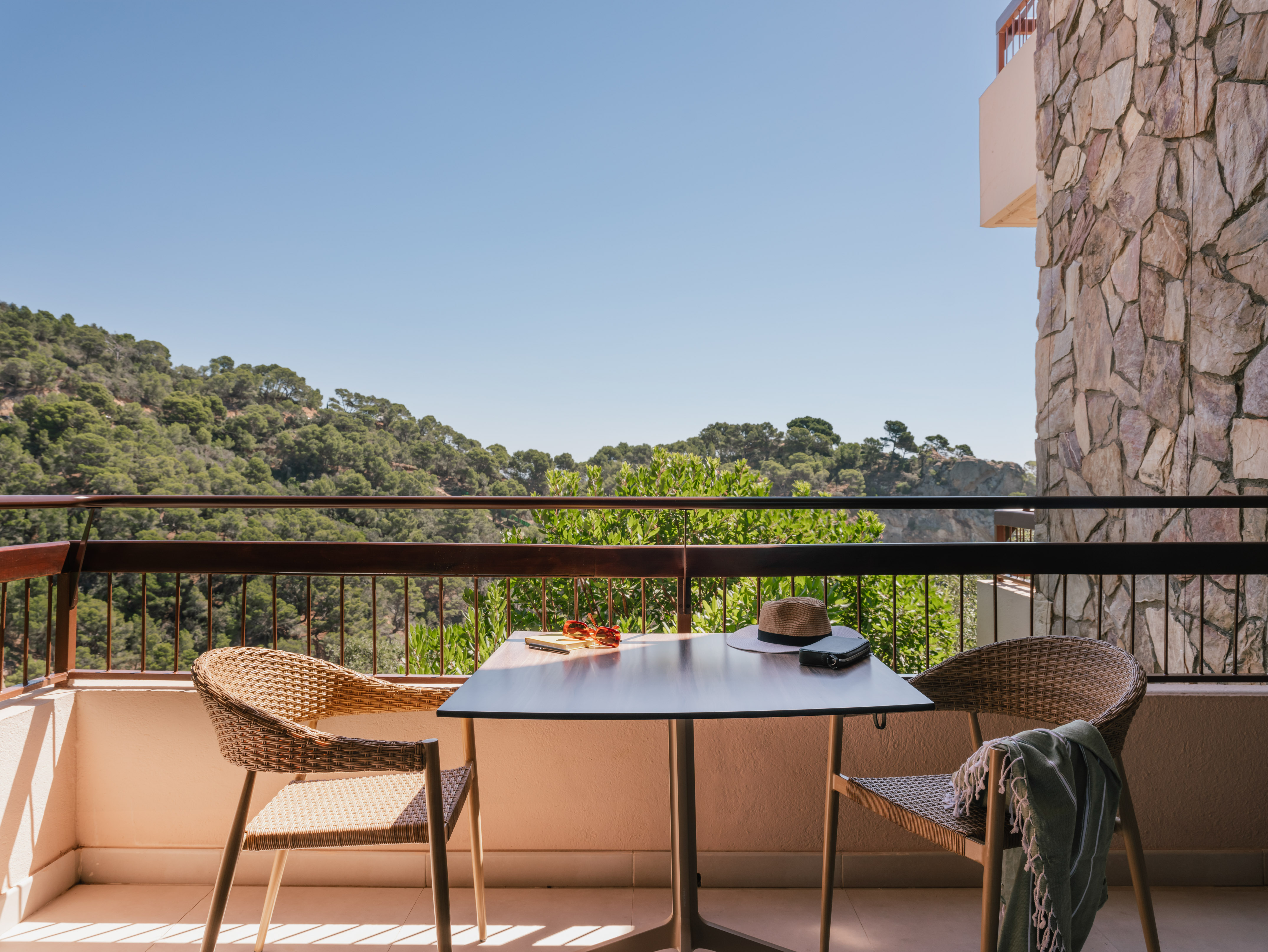 Sunny hotel balcony with wicker chairs, table, and forest mountain view.