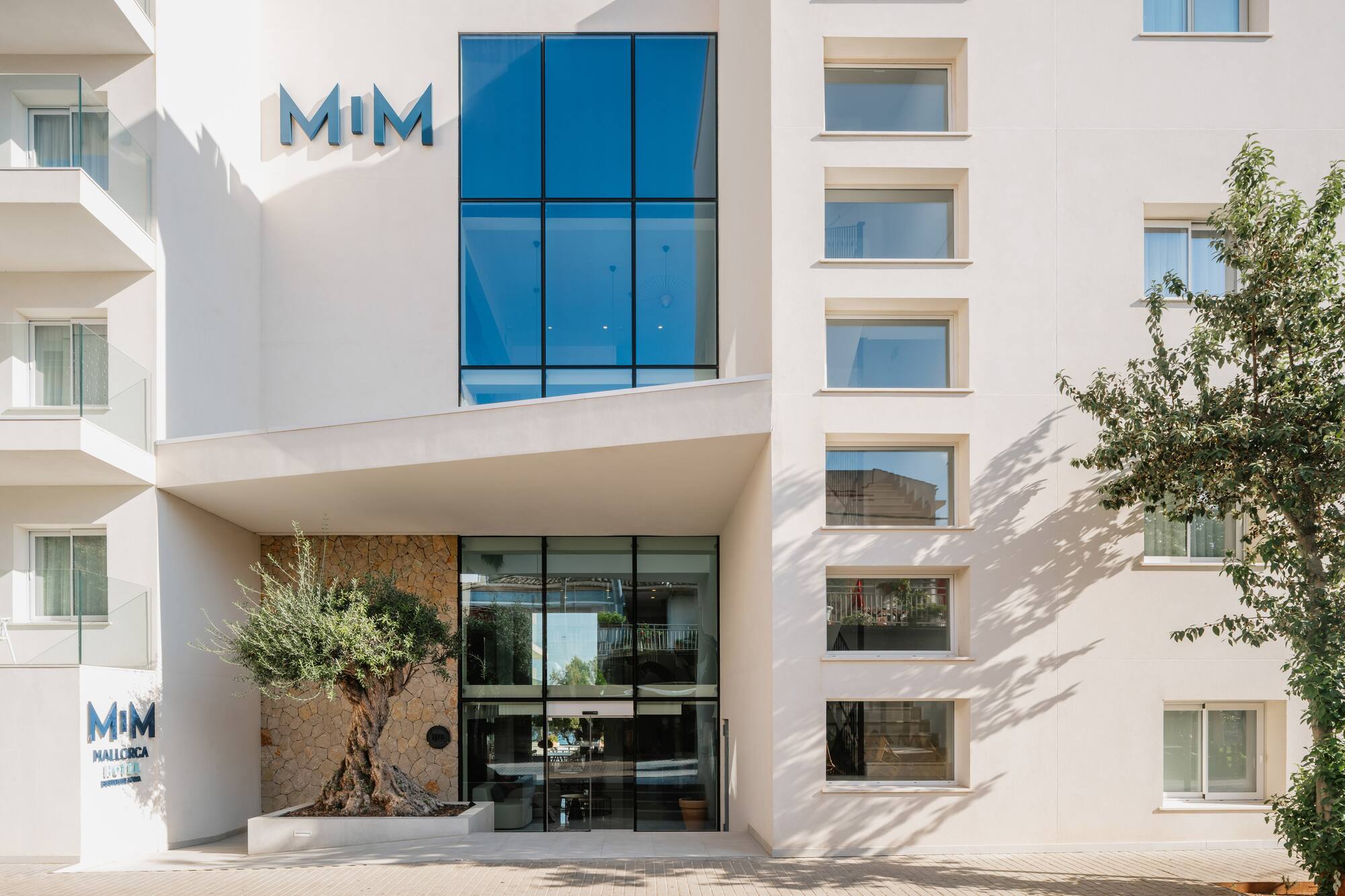 MIM Mallorca Hotel entrance with a modern white facade, glass, and olive tree.