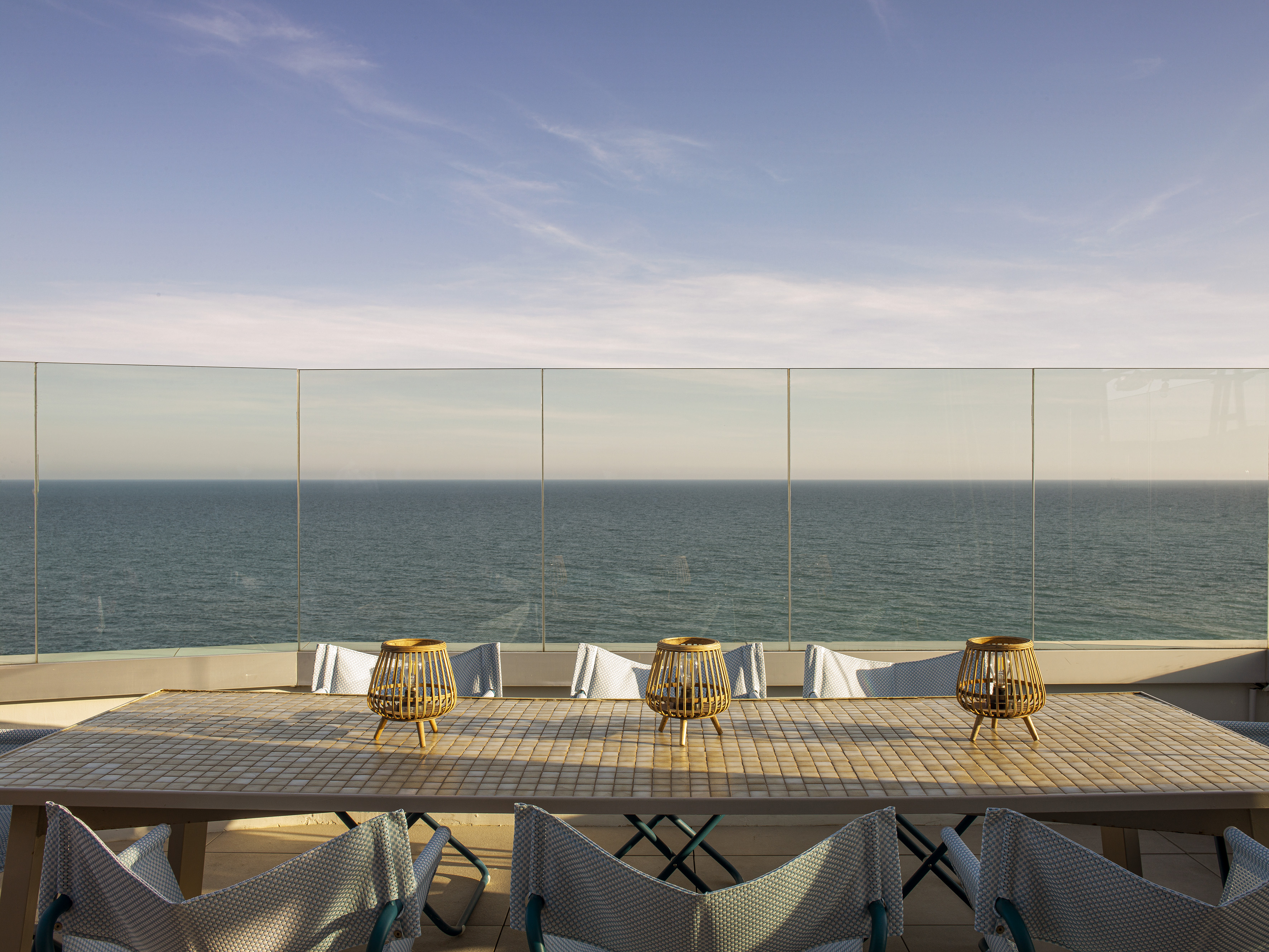 Coastal rooftop patio with dining table, lanterns, chairs, and ocean view.