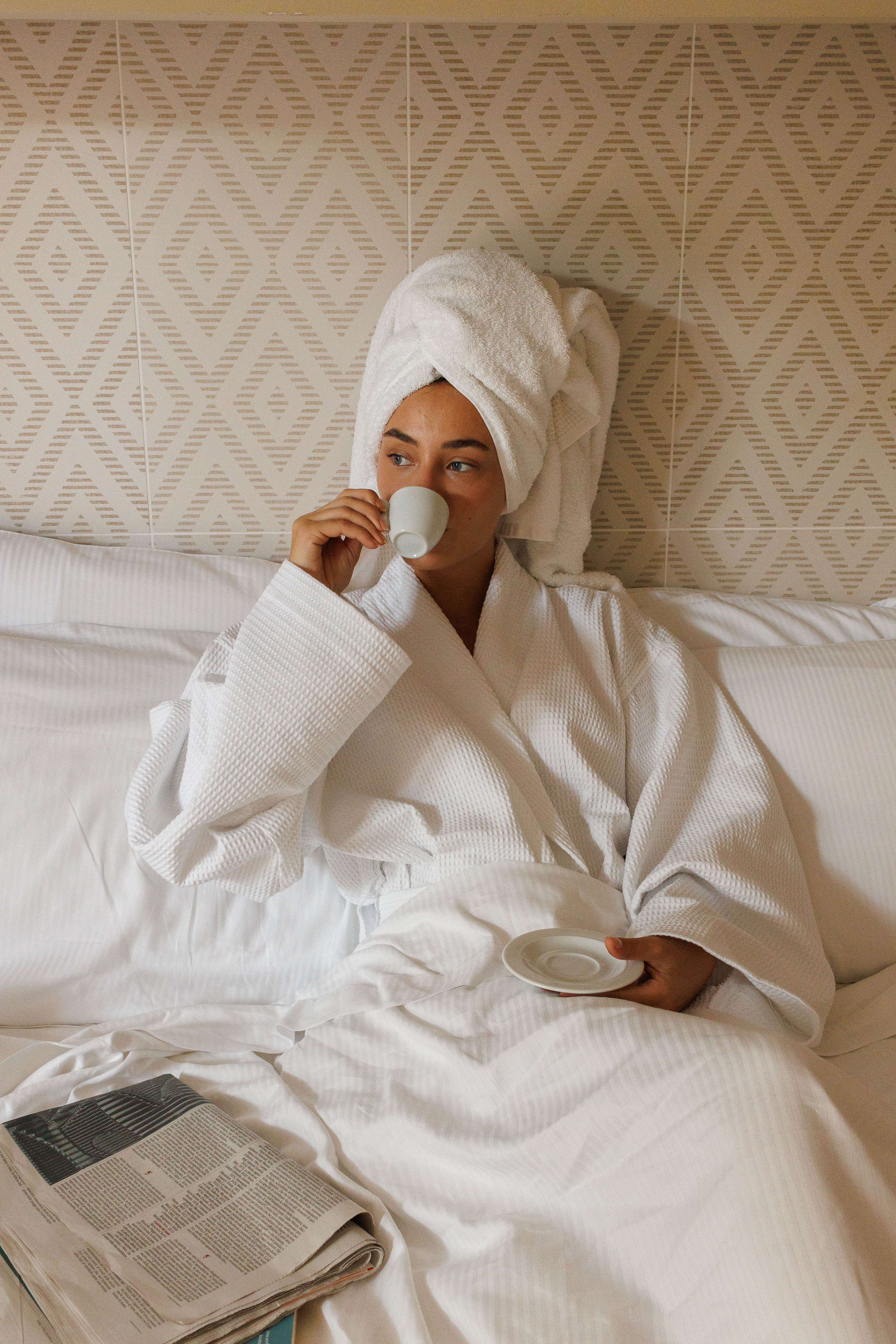 Woman in white bathrobe, towel on head, sips morning coffee in bed.