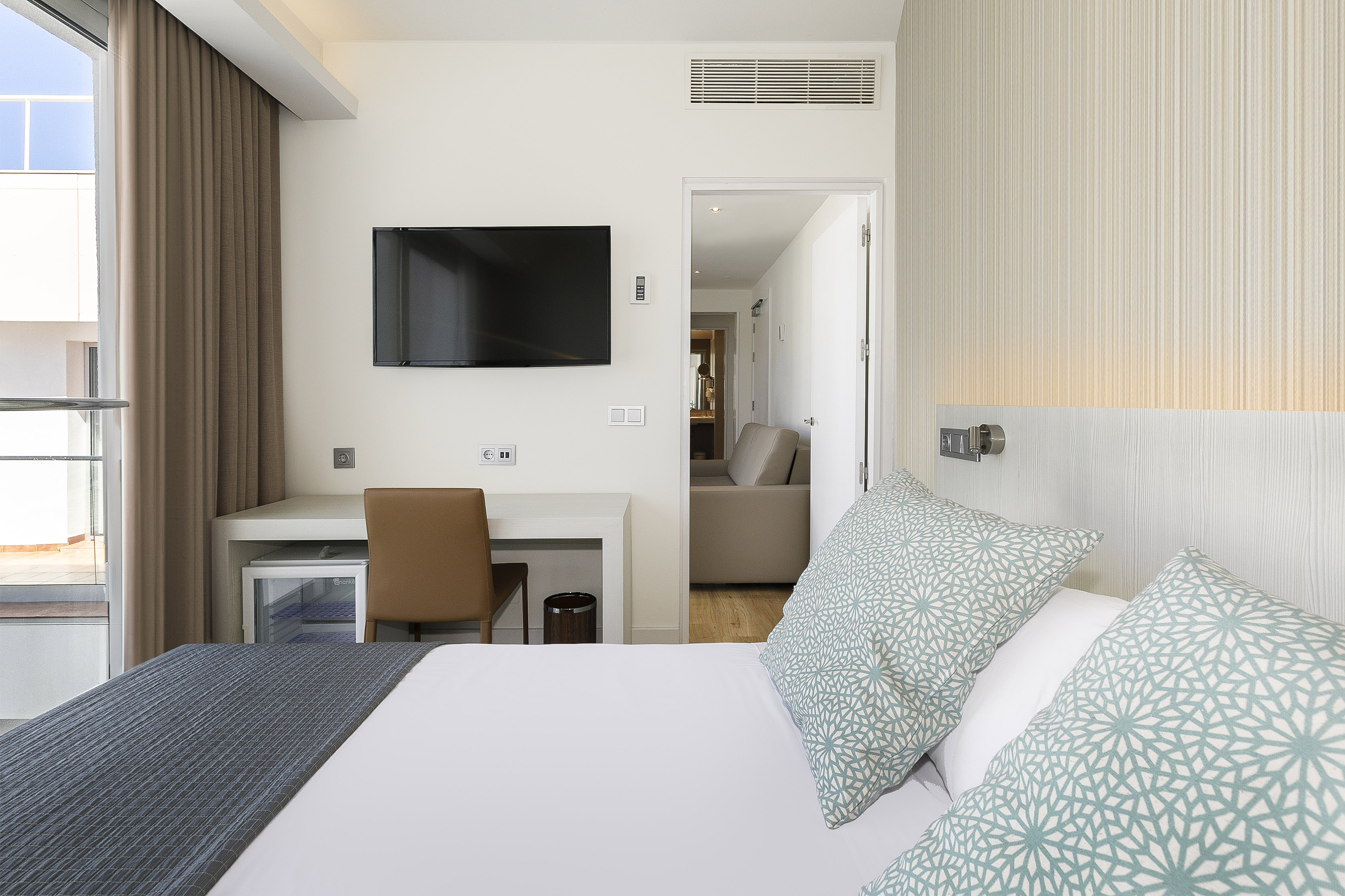 Modern hotel bedroom with bed, patterned pillows, desk, TV, and balcony.