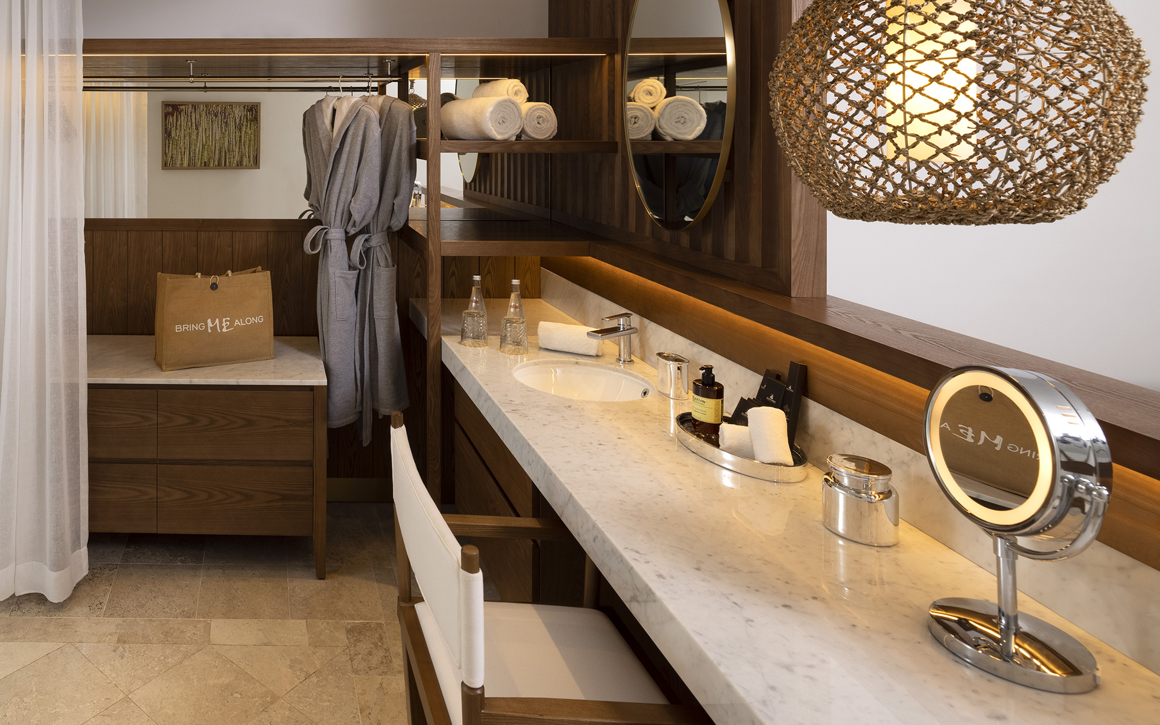 Modern hotel bathroom vanity, marble counter, wooden shelves, robes, makeup mirror.