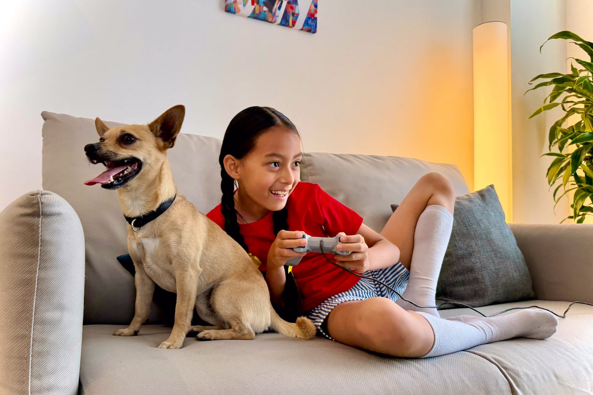 Smiling girl playing video games with a happy dog on a couch.