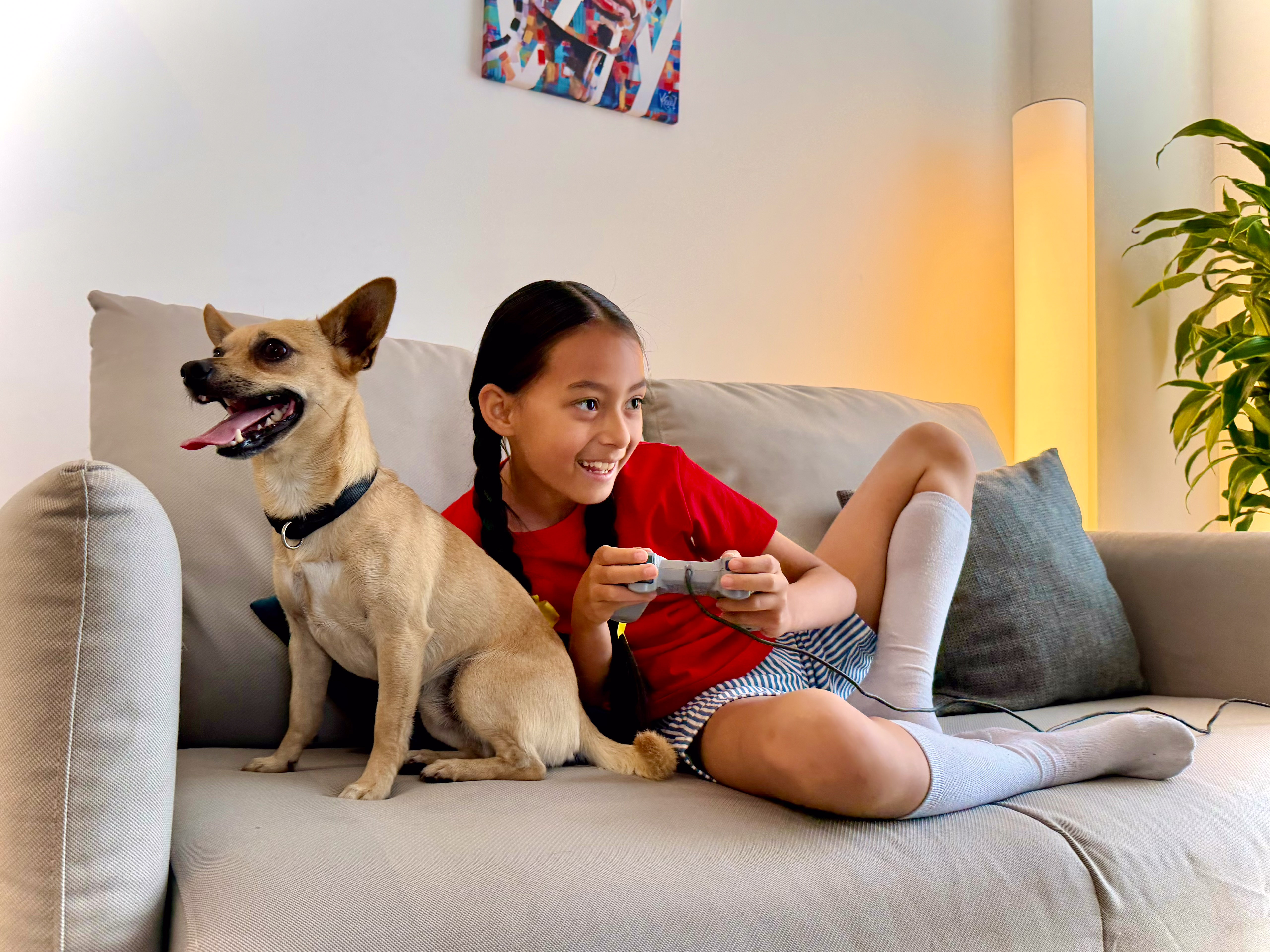 Smiling girl playing video games with a happy dog on a couch.