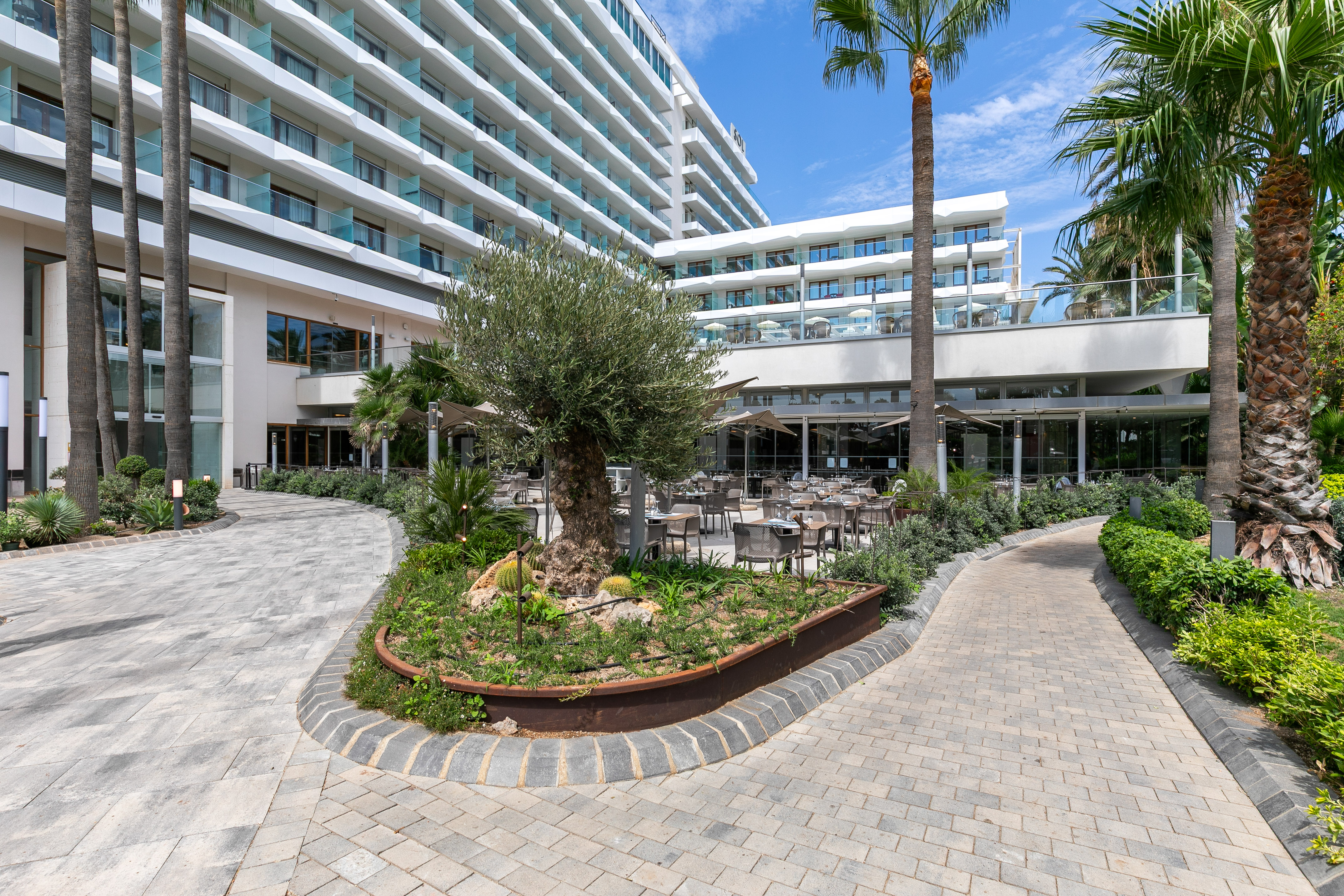 Modern hotel exterior with outdoor dining patio, palm trees, and paved paths.