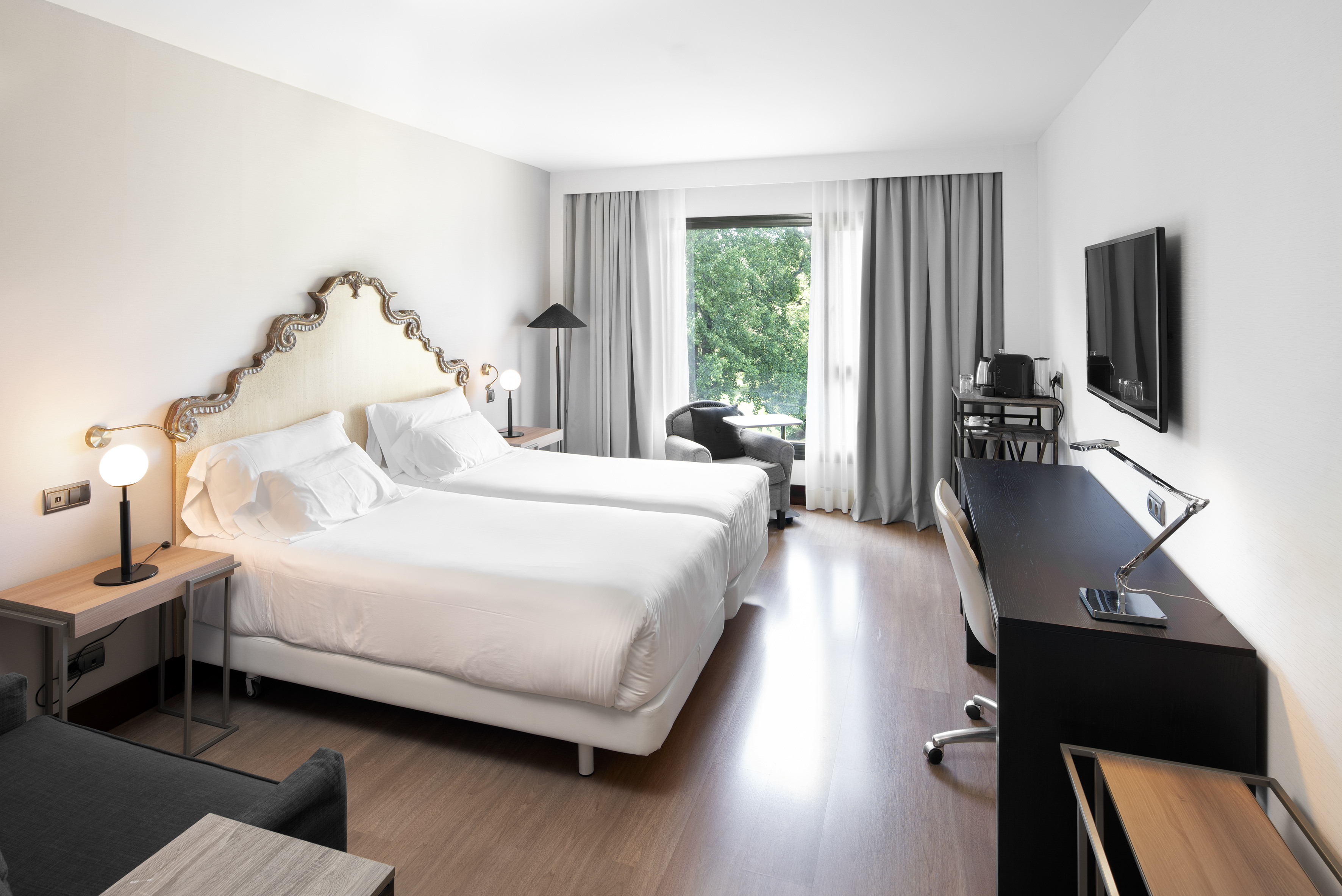 Elegant hotel room with twin beds, ornate headboard, desk, TV, and large window.