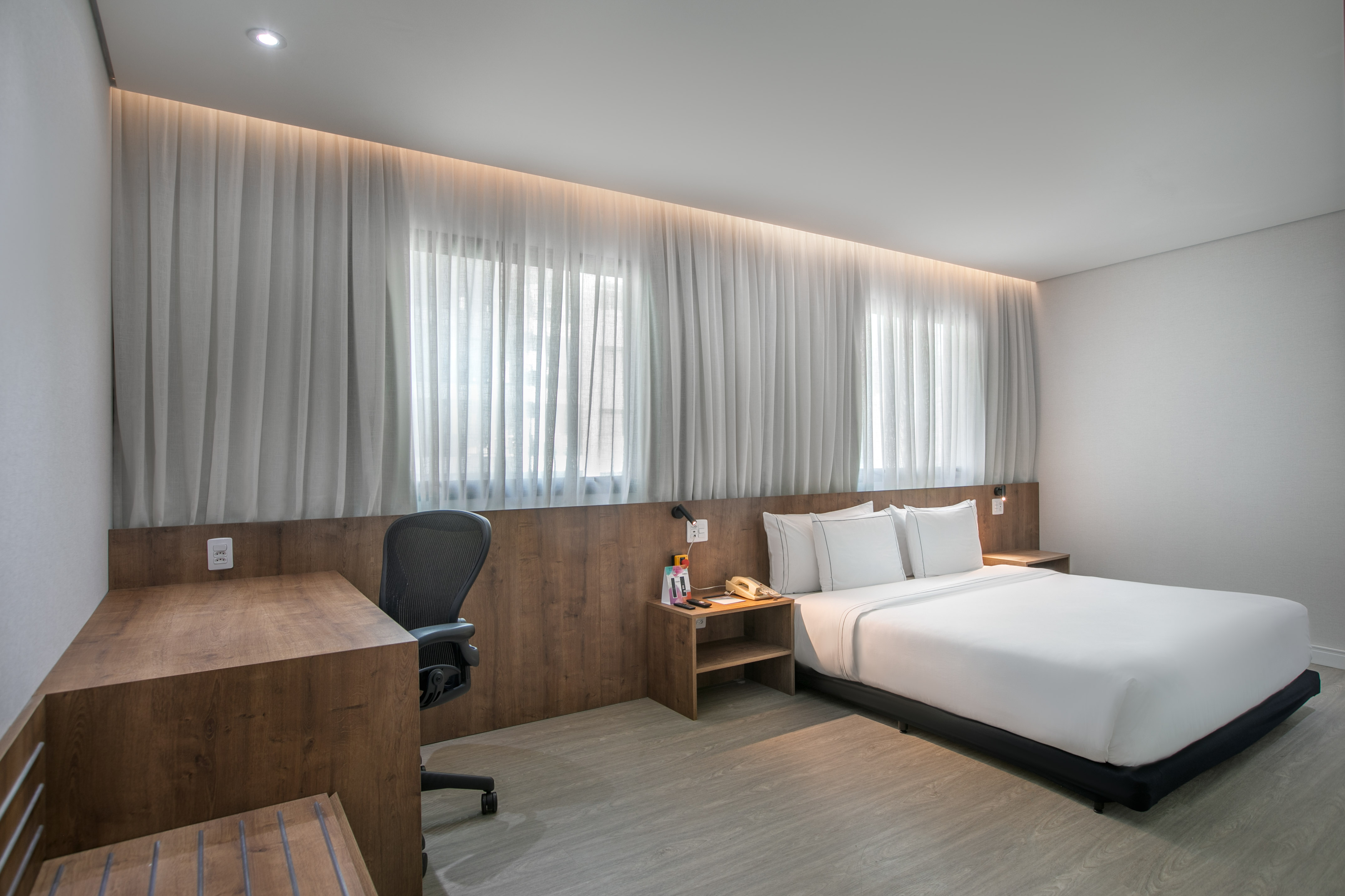Modern hotel guest room with queen bed, wood desk, and sheer window curtains.