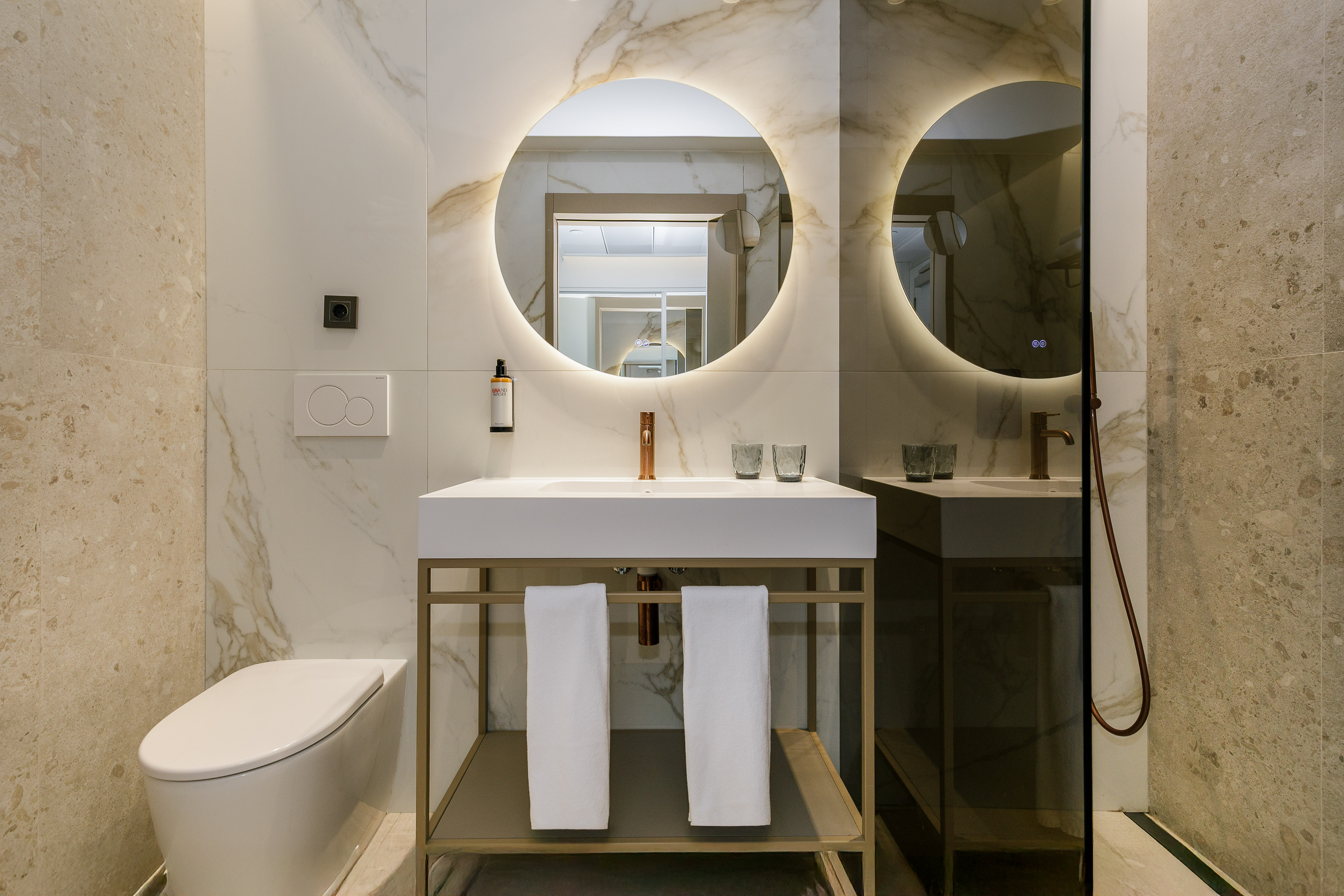 Modern bathroom with round illuminated mirror, marble walls, white vanity, and toilet.