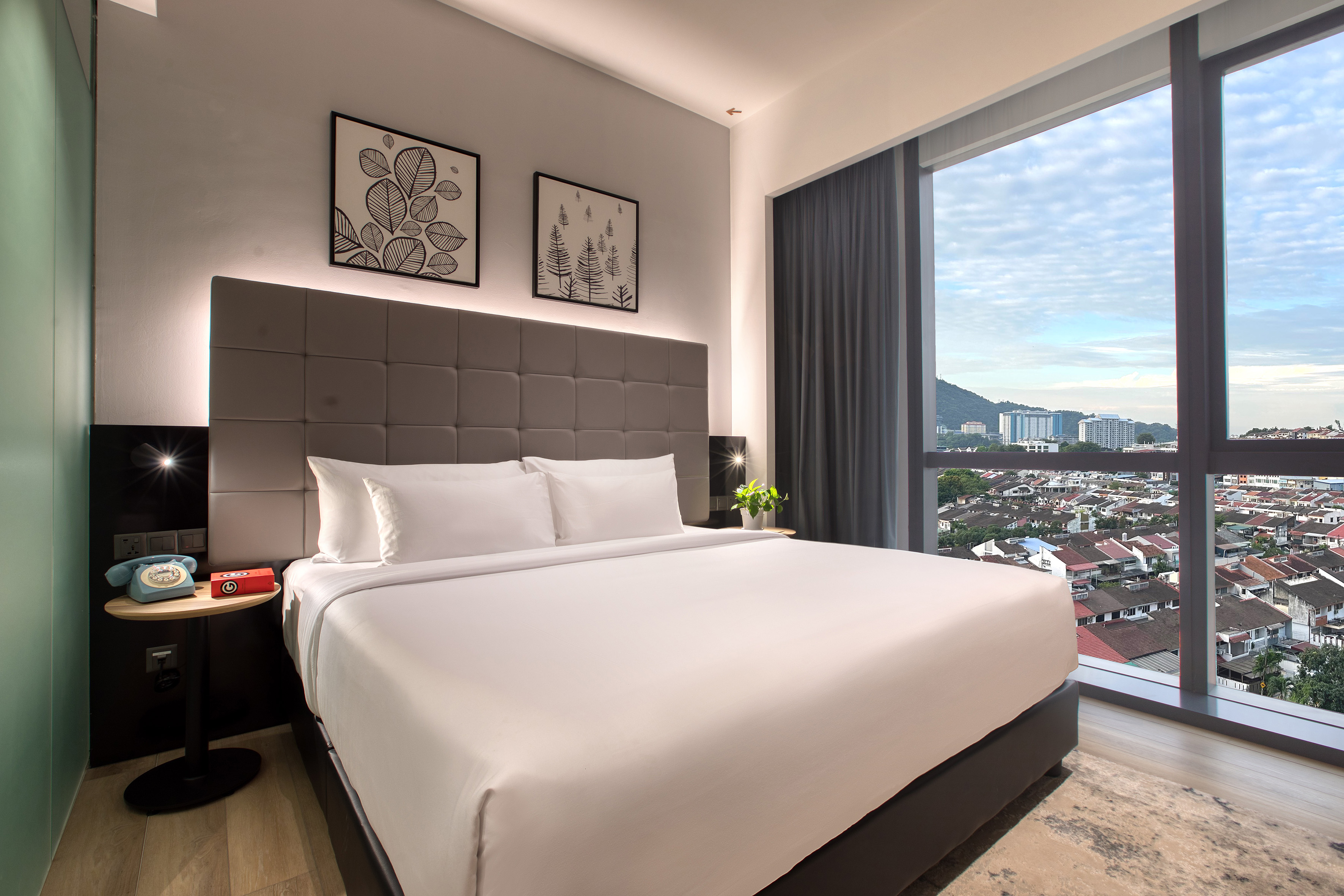 Modern hotel bedroom with white bed, city view through large window.