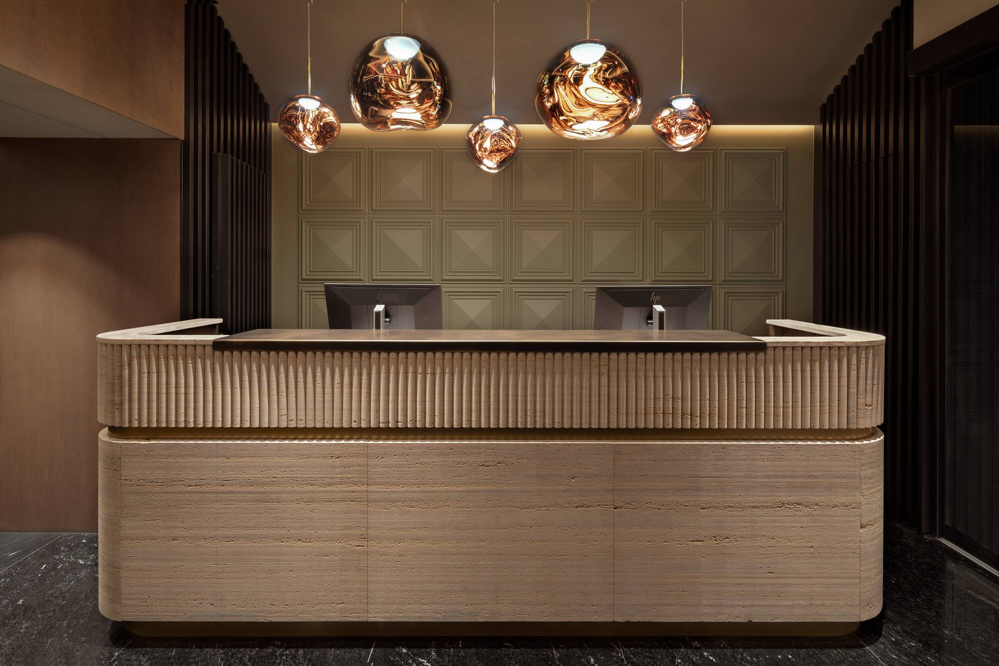 Modern hotel reception desk with ribbed travertine, geometric wall, copper pendant lights.