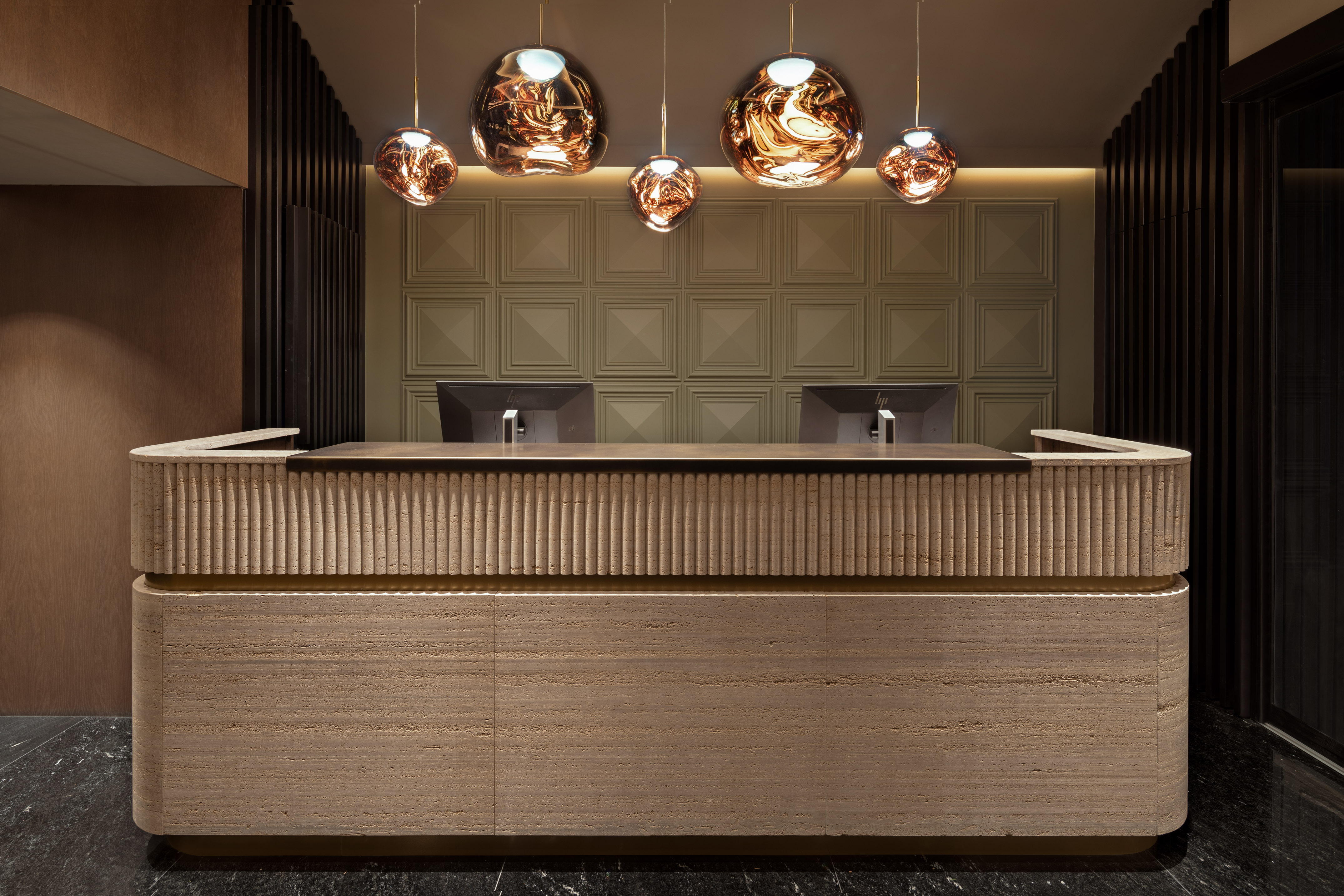 Modern hotel reception desk with ribbed travertine, geometric wall, copper pendant lights.