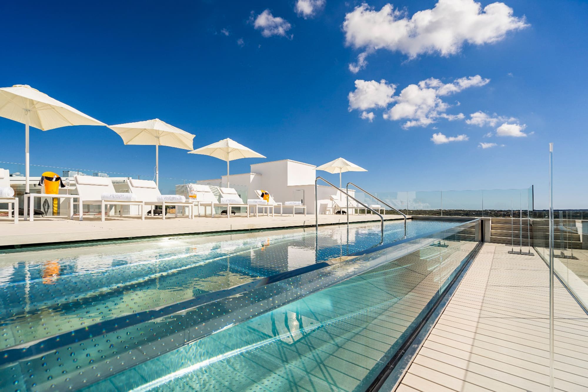 Rooftop infinity pool, transparent sides, white lounge chairs, sunny day.