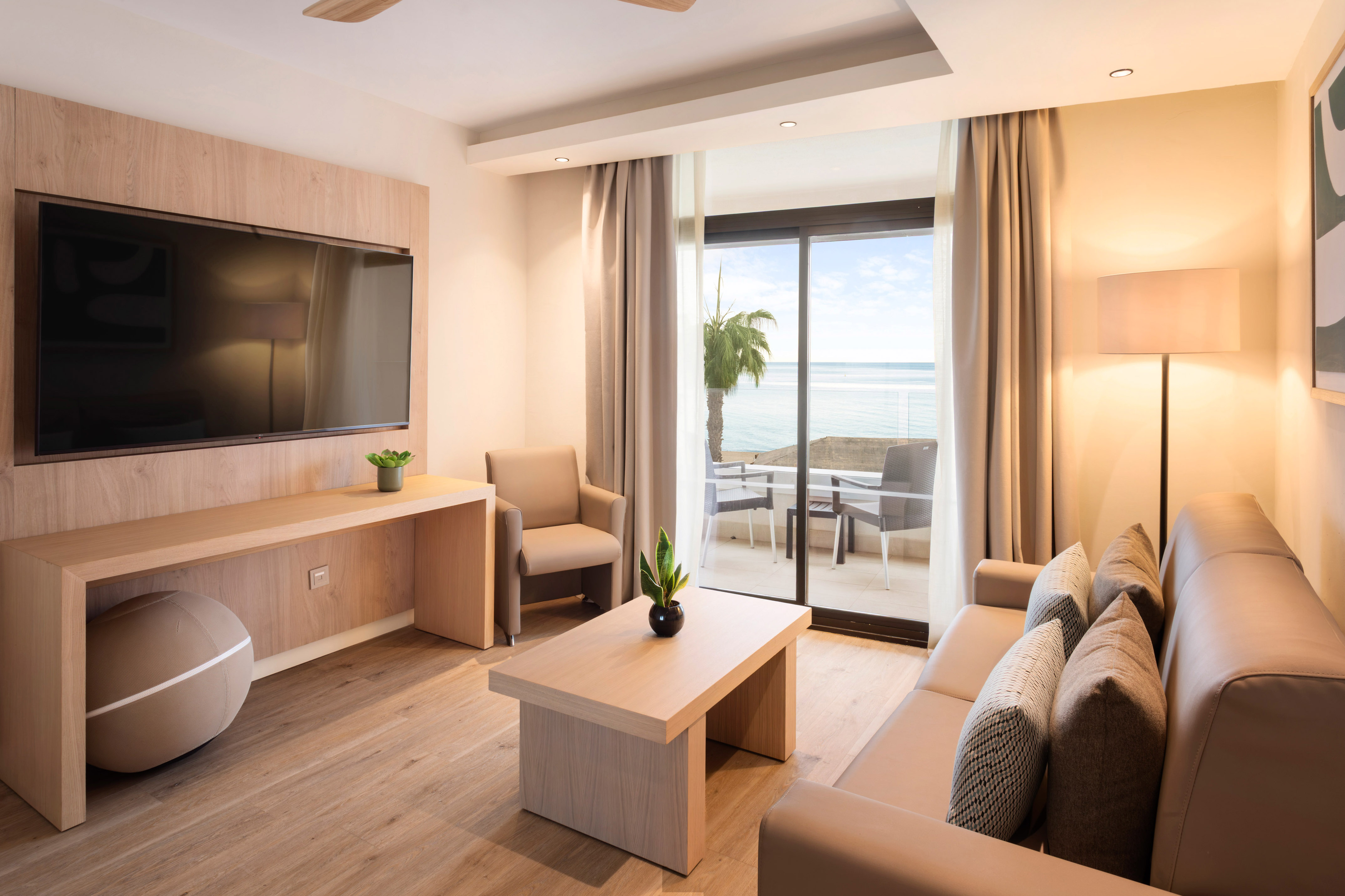 Modern hotel suite living room with ocean view, balcony, and large TV.