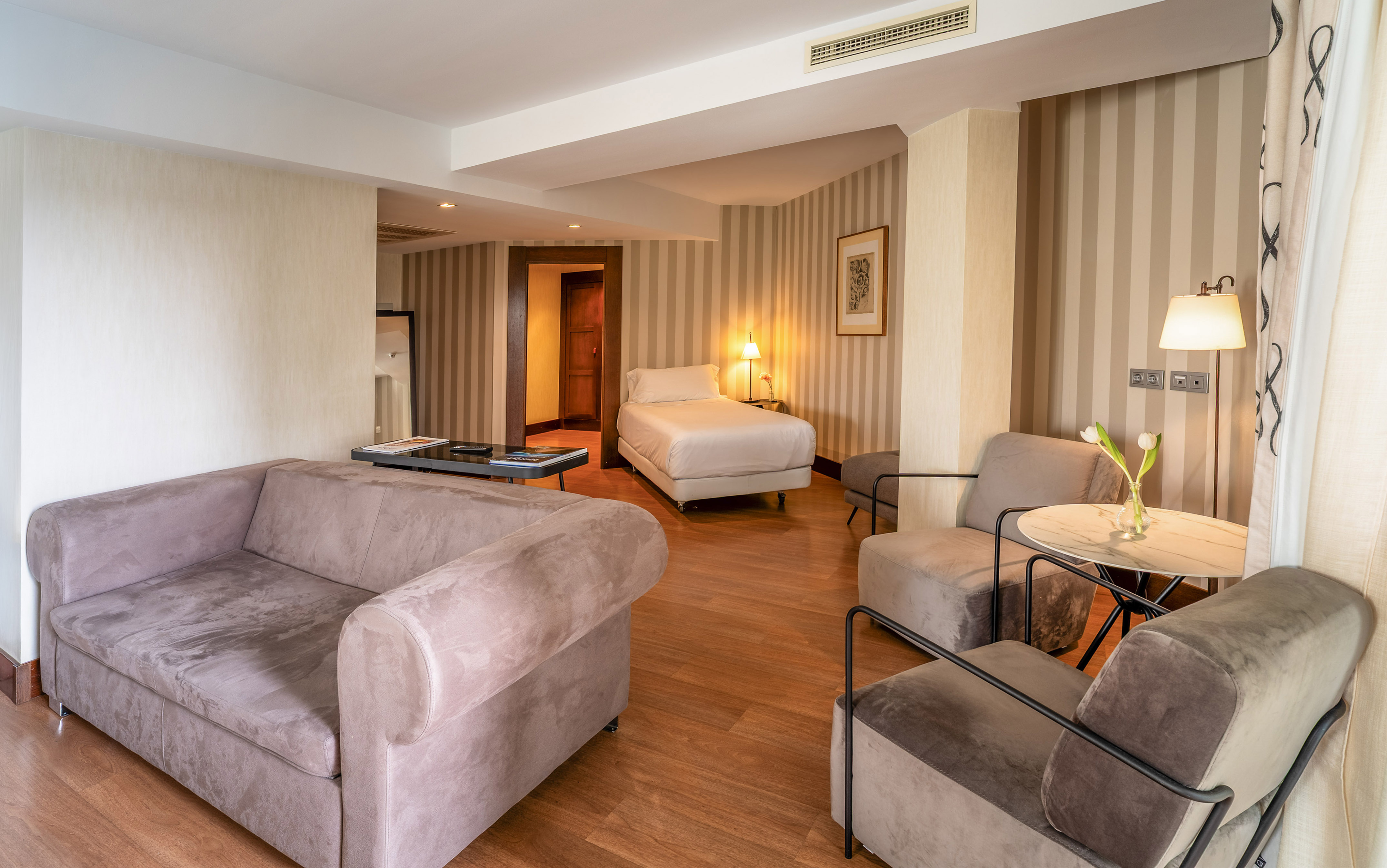 Modern hotel suite with living area, single bed, sofa, and hardwood floor.