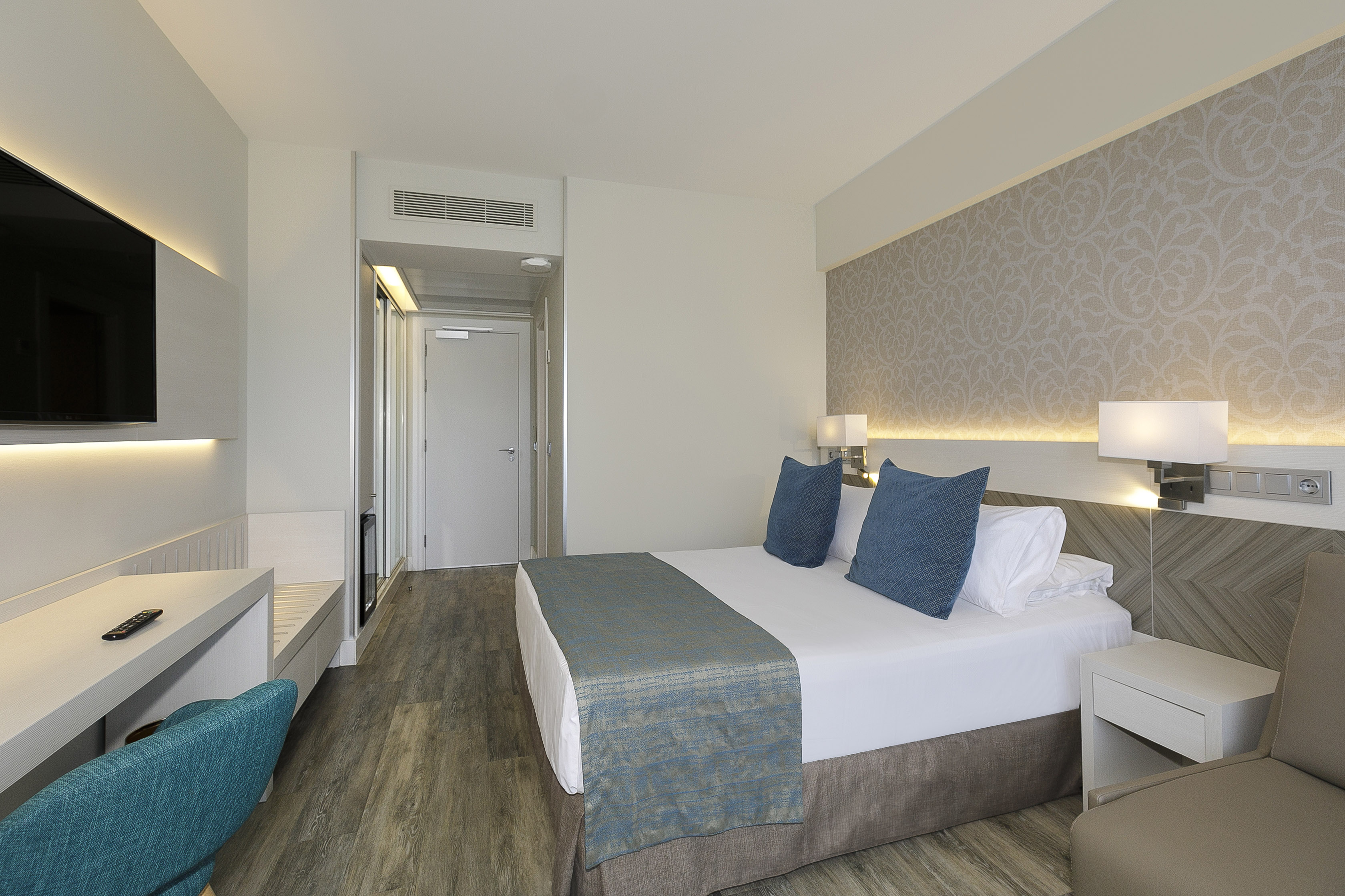 Modern hotel room interior with queen bed, blue pillows, patterned wall, and desk.