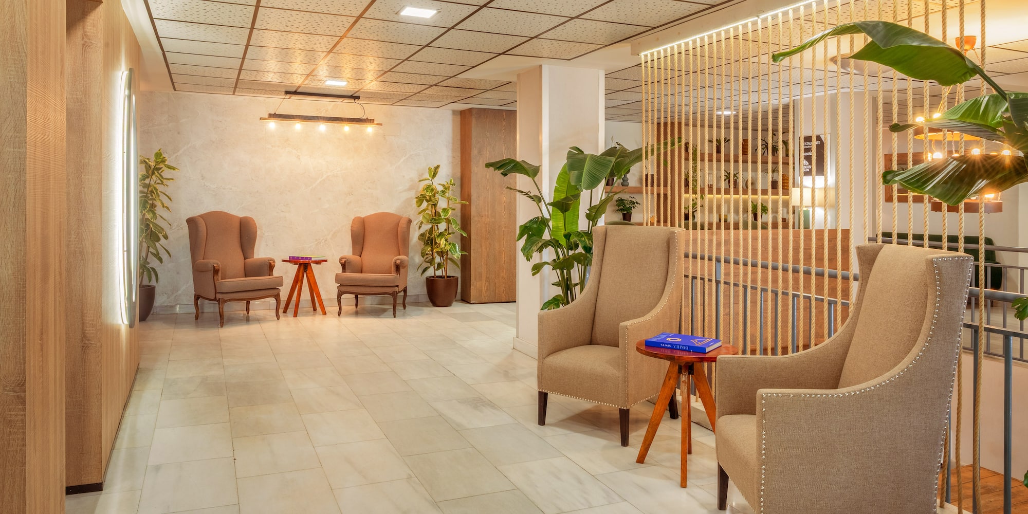 Modern lobby with beige armchairs, rope partition, marble floor, and plants.