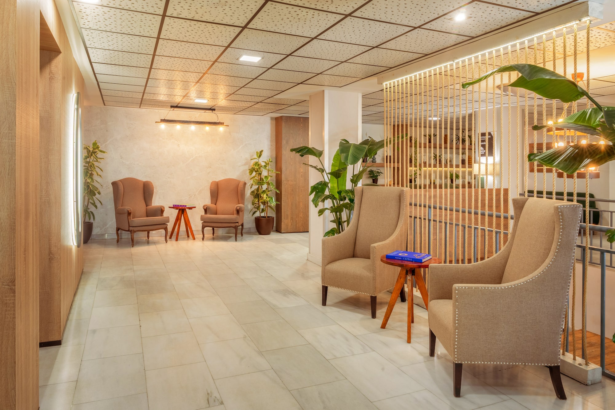 Modern lobby with beige armchairs, rope partition, marble floor, and plants.