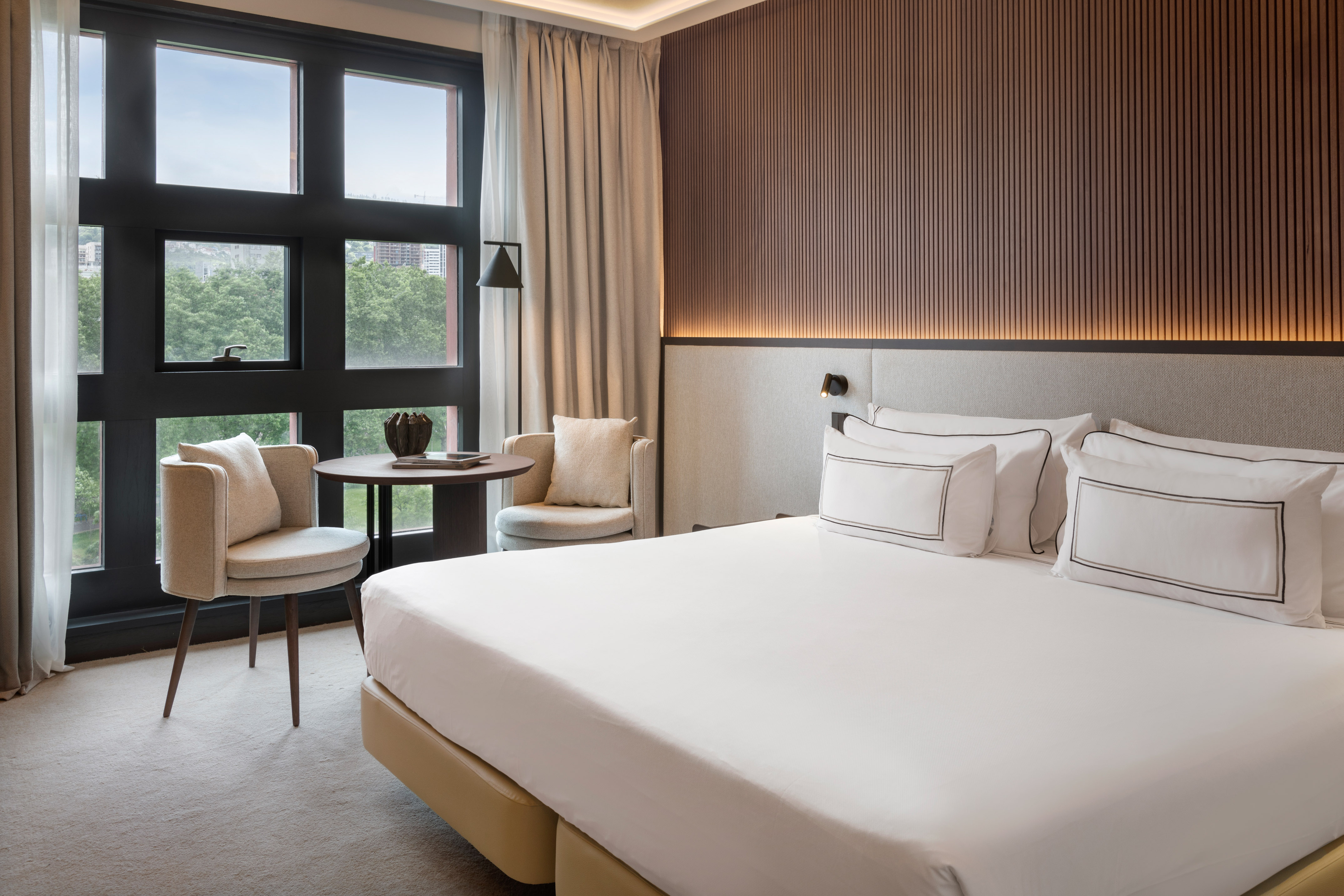 Modern hotel room with bed, large window, two chairs, and wood accent wall.