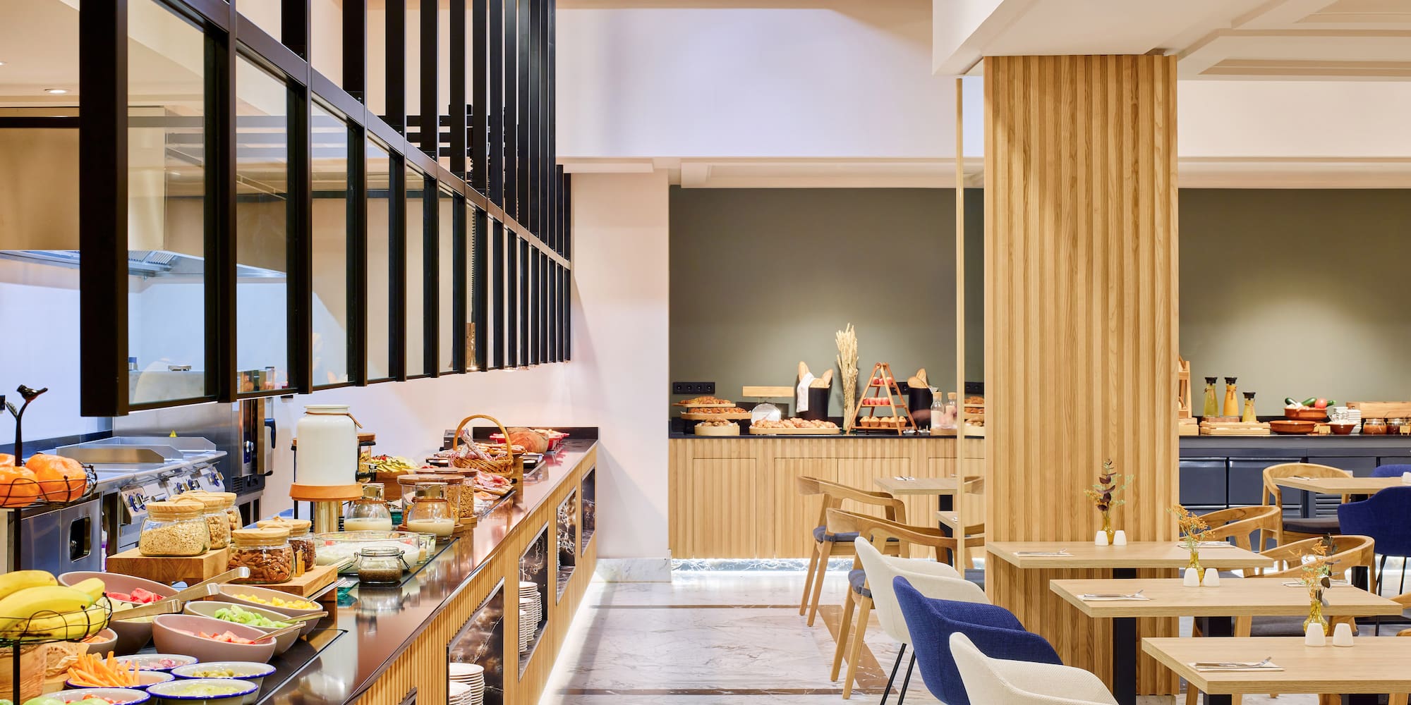 Buffet de desayuno moderno en hotel, con frutas, cereales y zona de mesas.