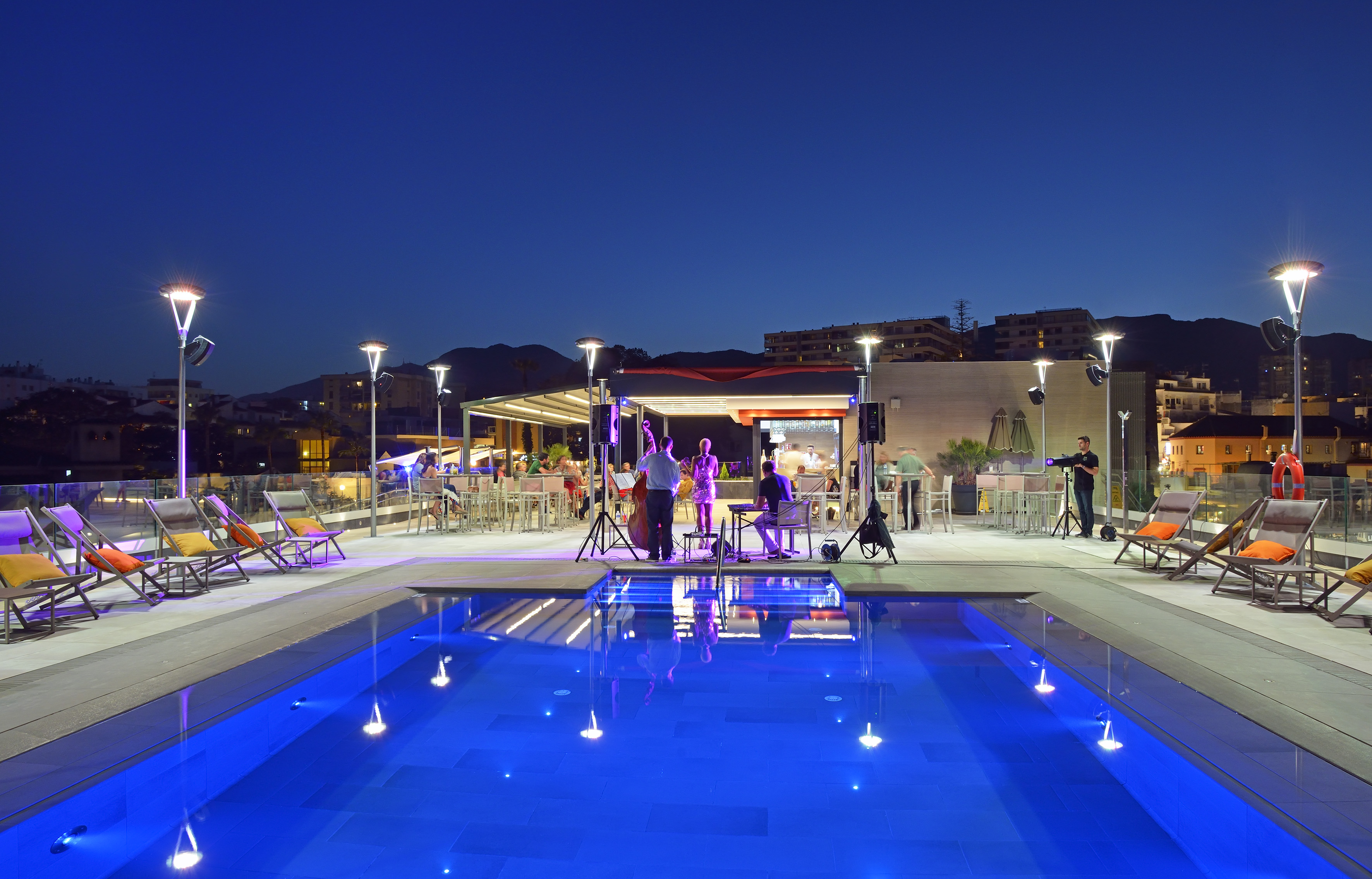 Illuminated rooftop pool and bar with live music at night.