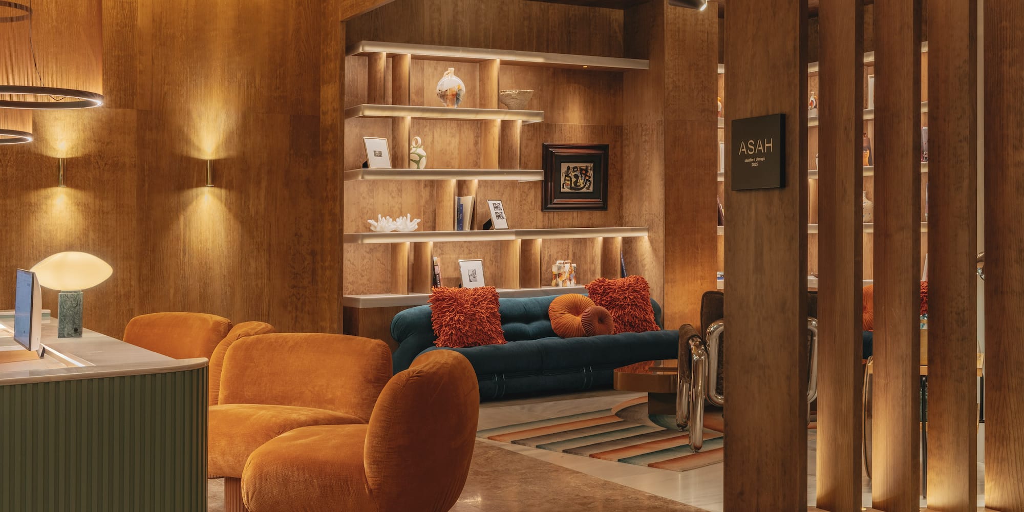 Modern wood-paneled lounge with warm lighting, orange chairs, and a blue sofa.