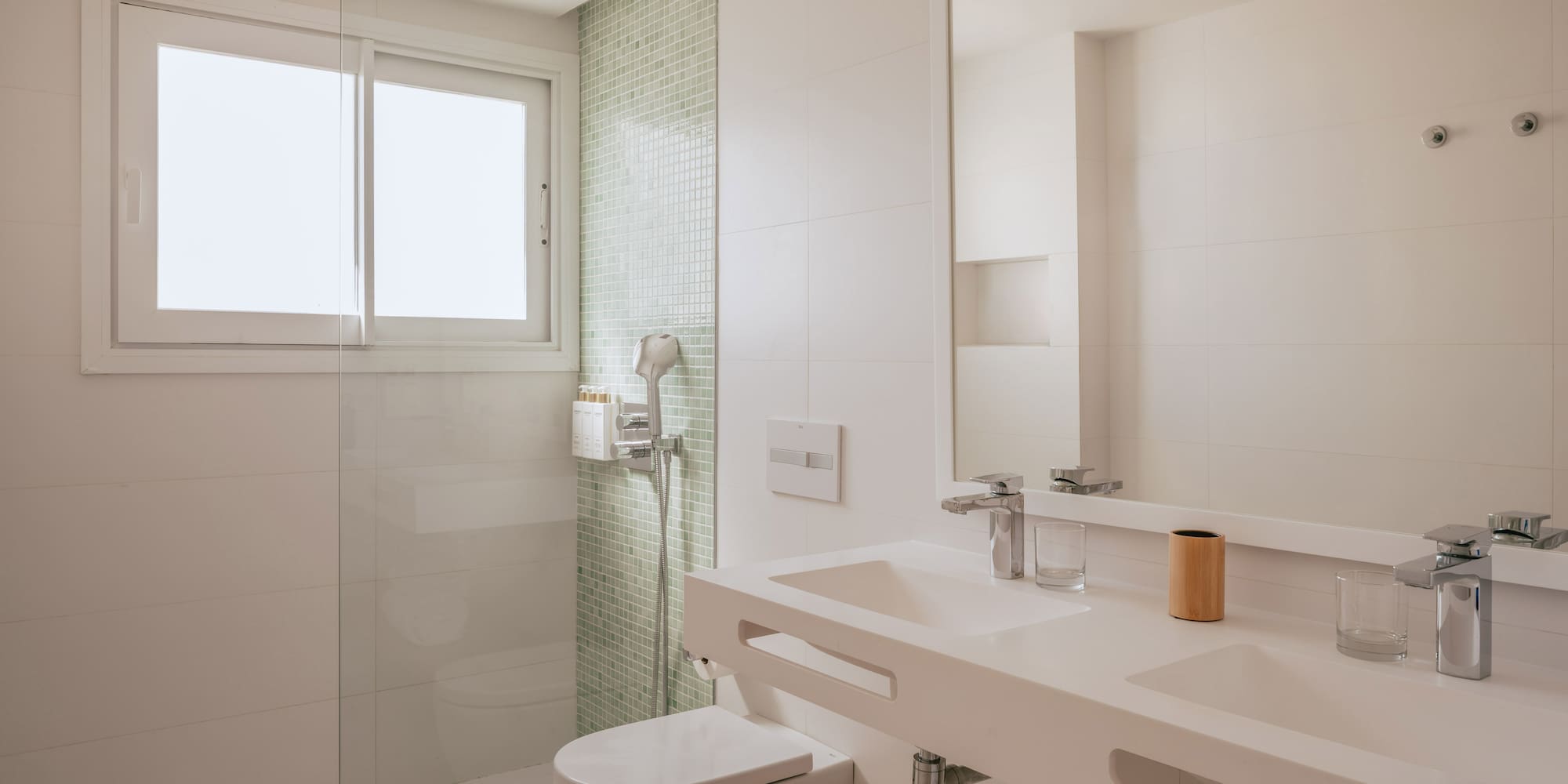 Modern white bathroom with glass shower, toilet, double vanity, and window.