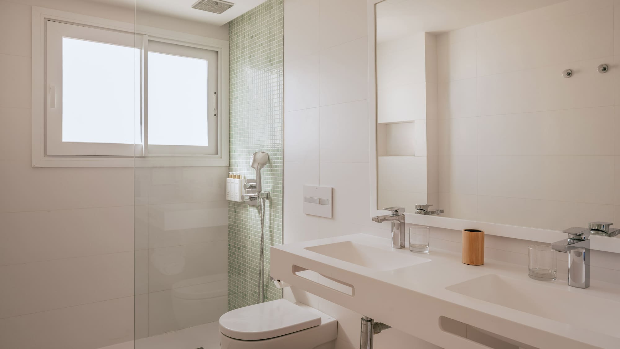 Modern white bathroom with glass shower, toilet, double vanity, and window.