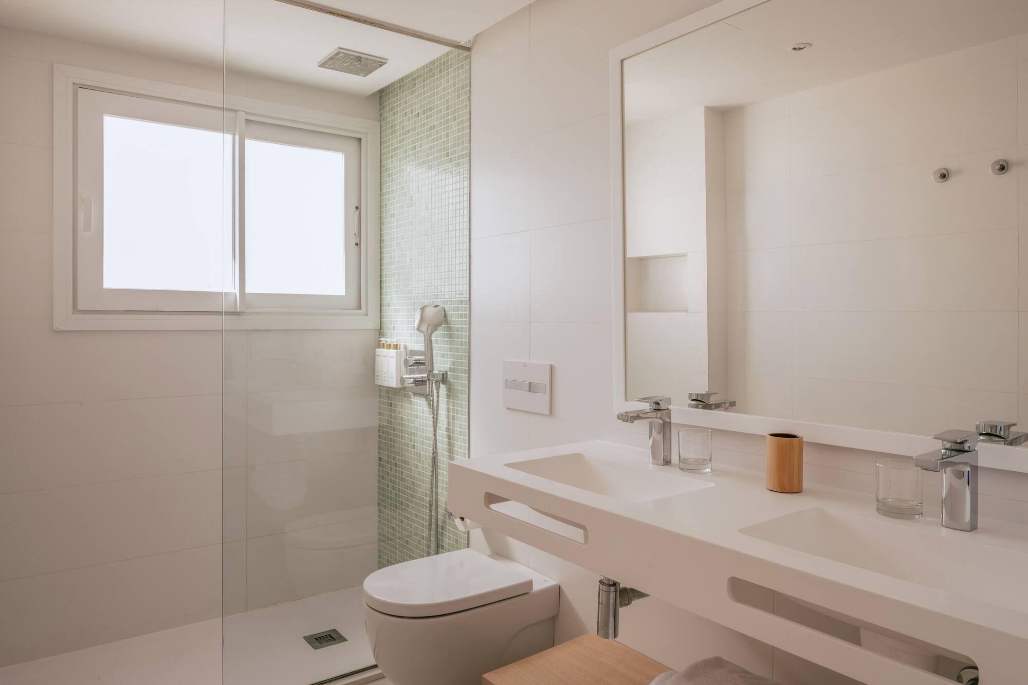 Baño moderno y luminoso con ducha de cristal, inodoro y doble lavabo. Azulejos blancos.