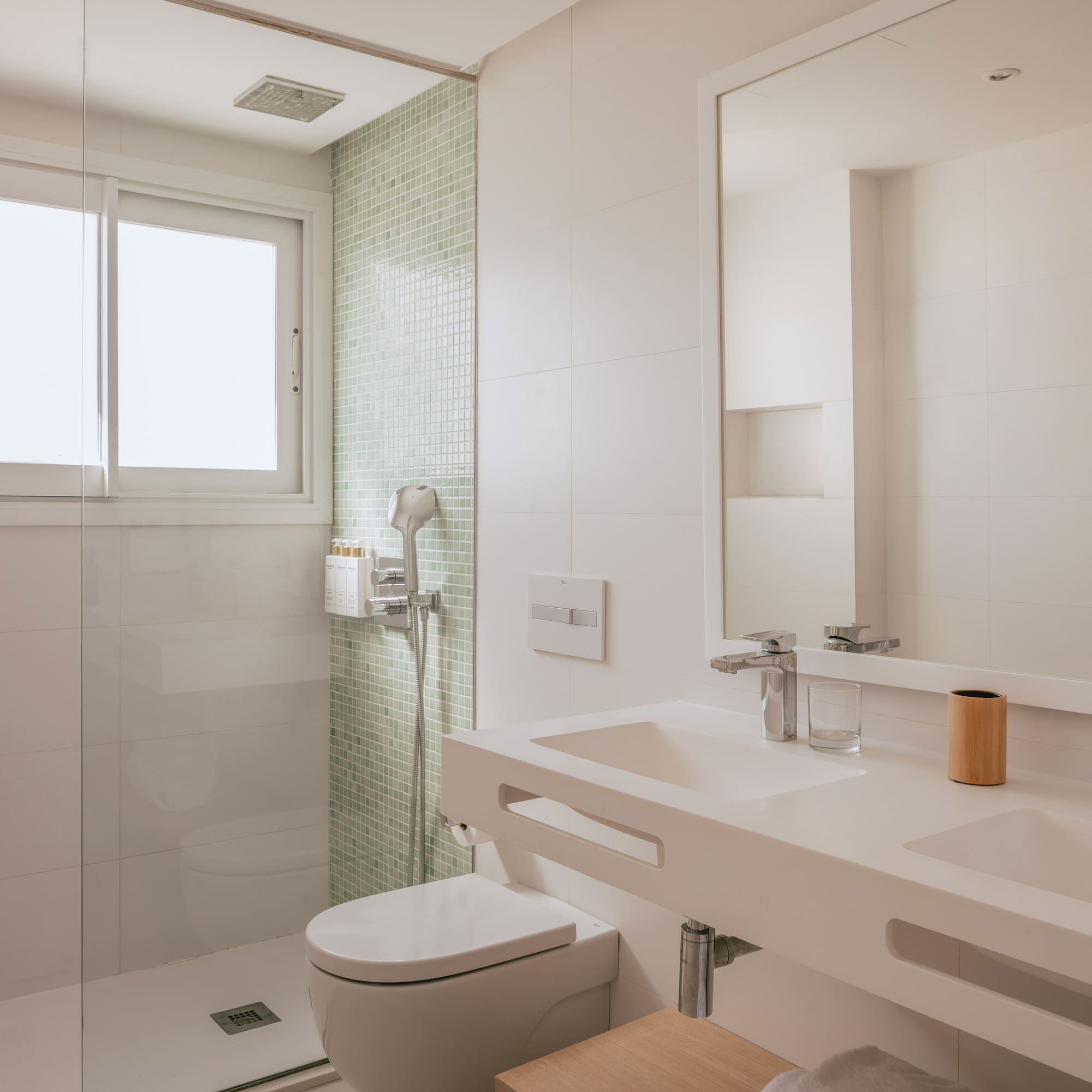 Modern white bathroom with glass shower, toilet, double vanity, and window.