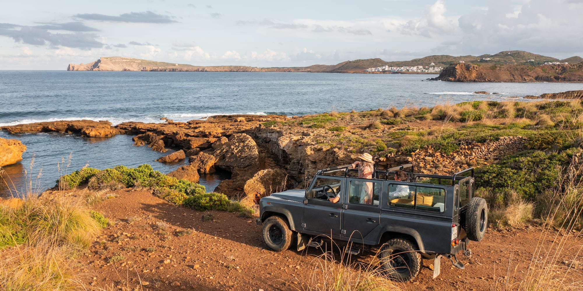 Grey 4x4 on a rocky coastal track, Mediterranean sea, distant village.