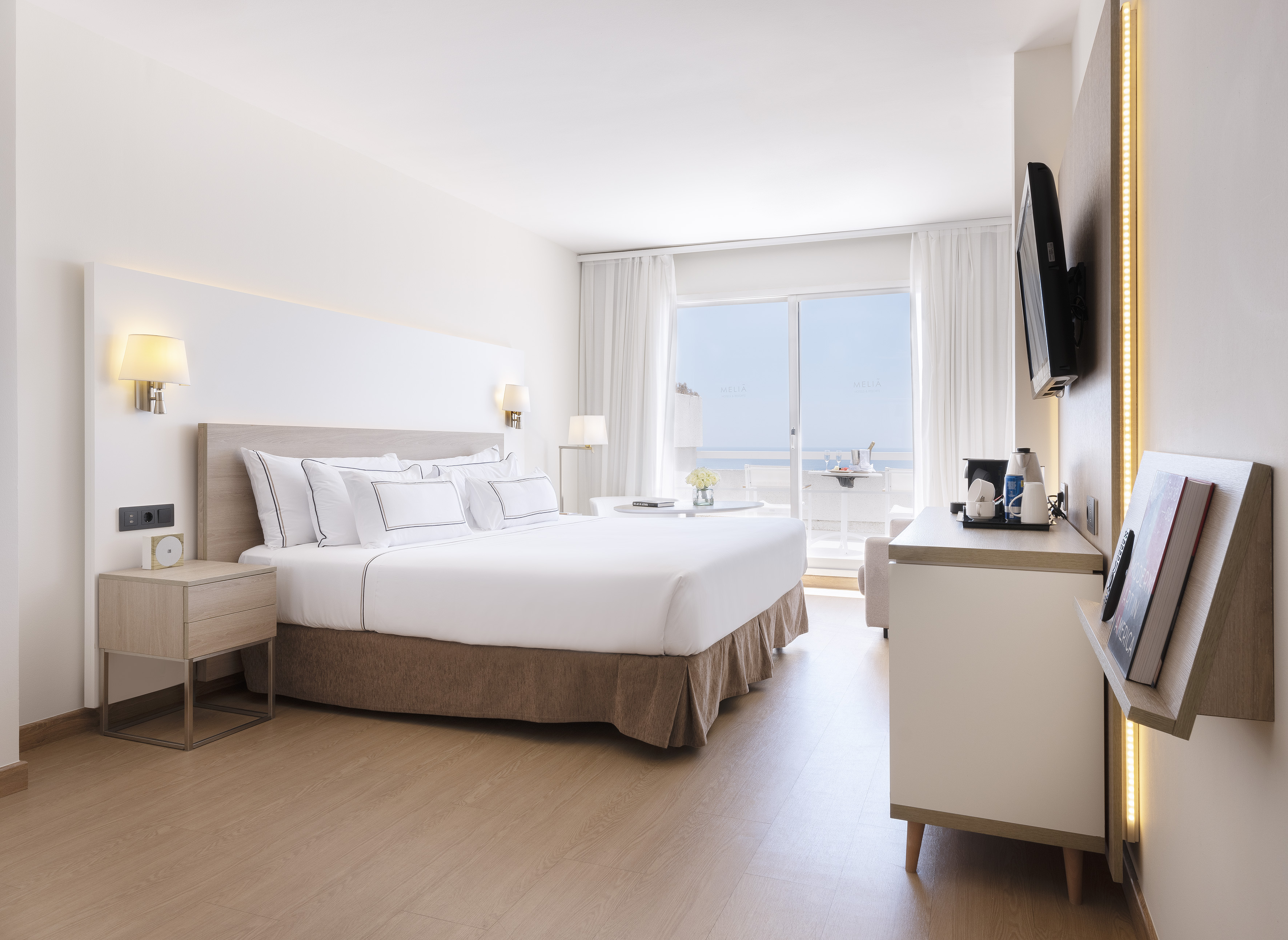 Modern hotel room with white bed, ocean view balcony, and light wood decor.