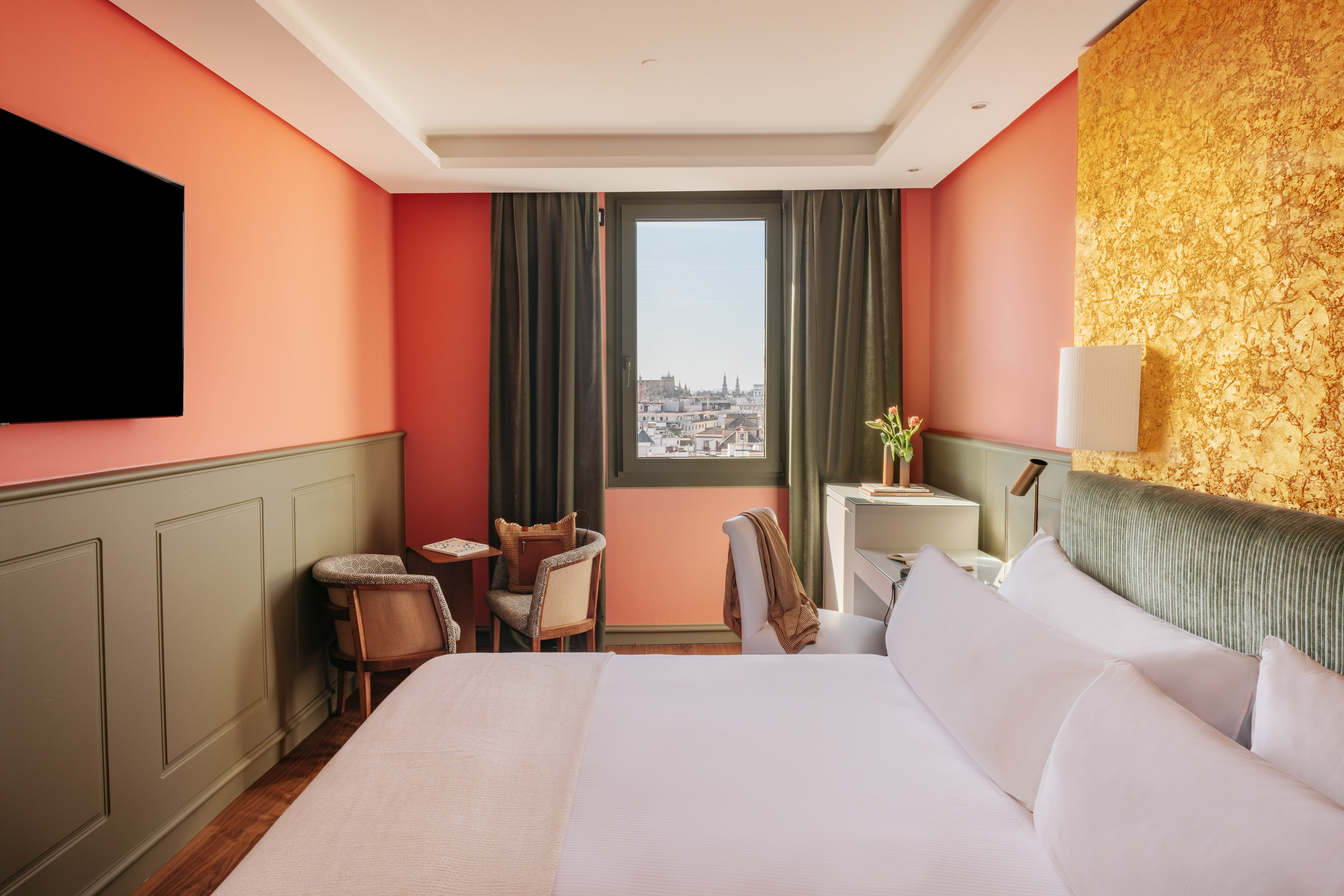 Modern hotel bedroom with coral walls, gold accent, large bed, city view.