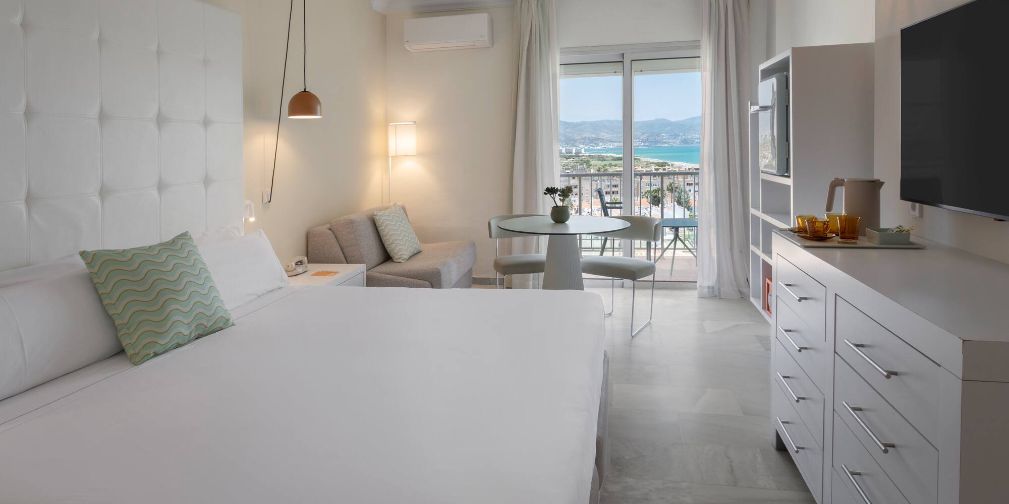 Suite de hotel luminosa con balcón, vistas al mar, cama doble y zona de estar.