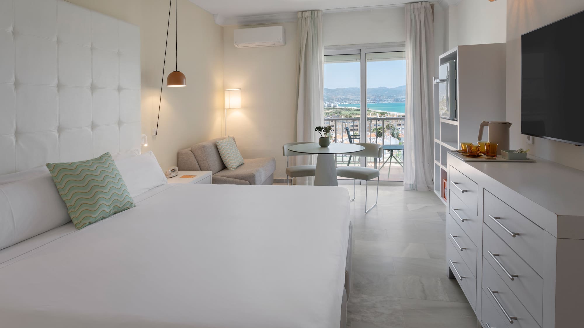 Suite de hotel luminosa con balcón, vistas al mar, cama doble y zona de estar.