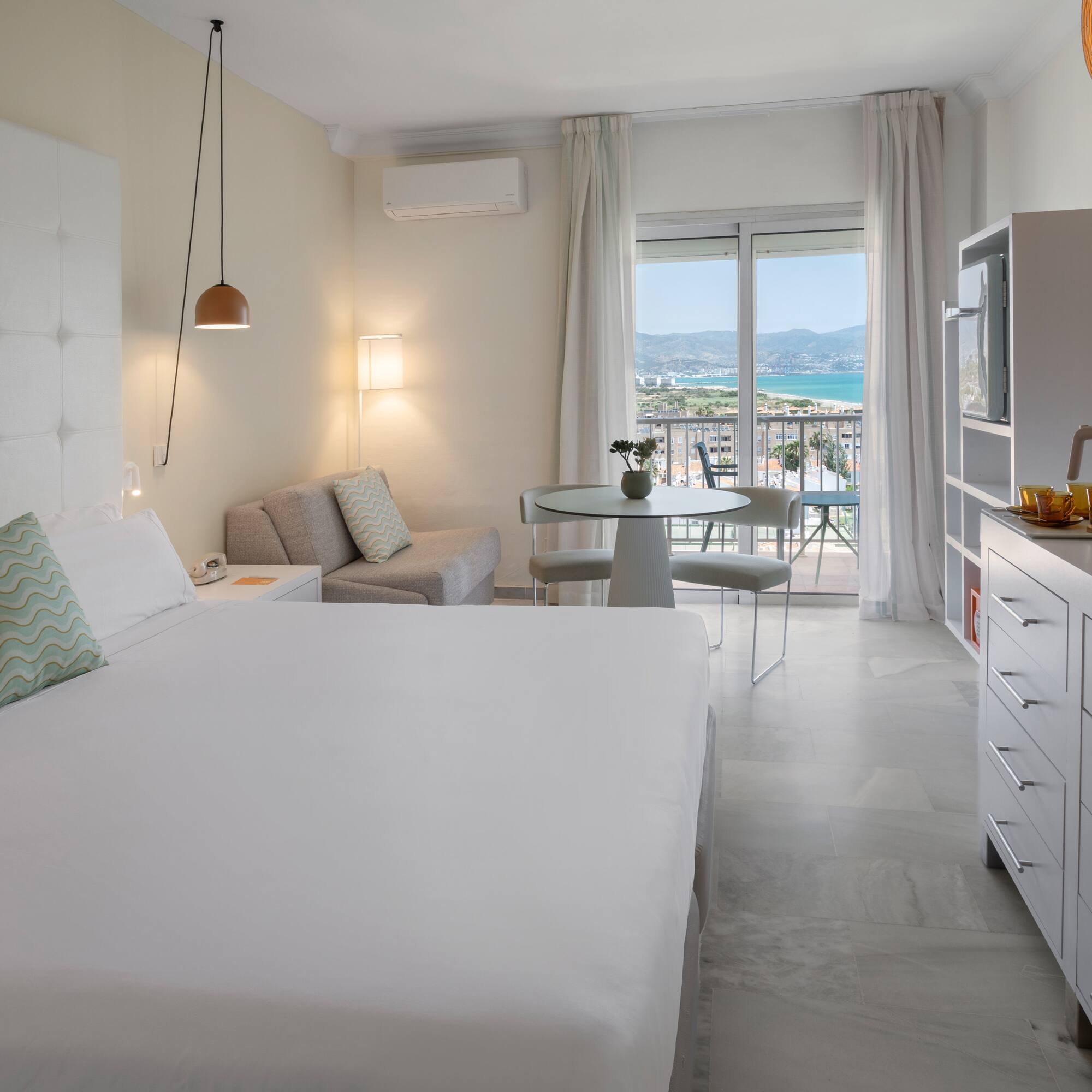Suite de hotel luminosa con balcón, vistas al mar, cama doble y zona de estar.