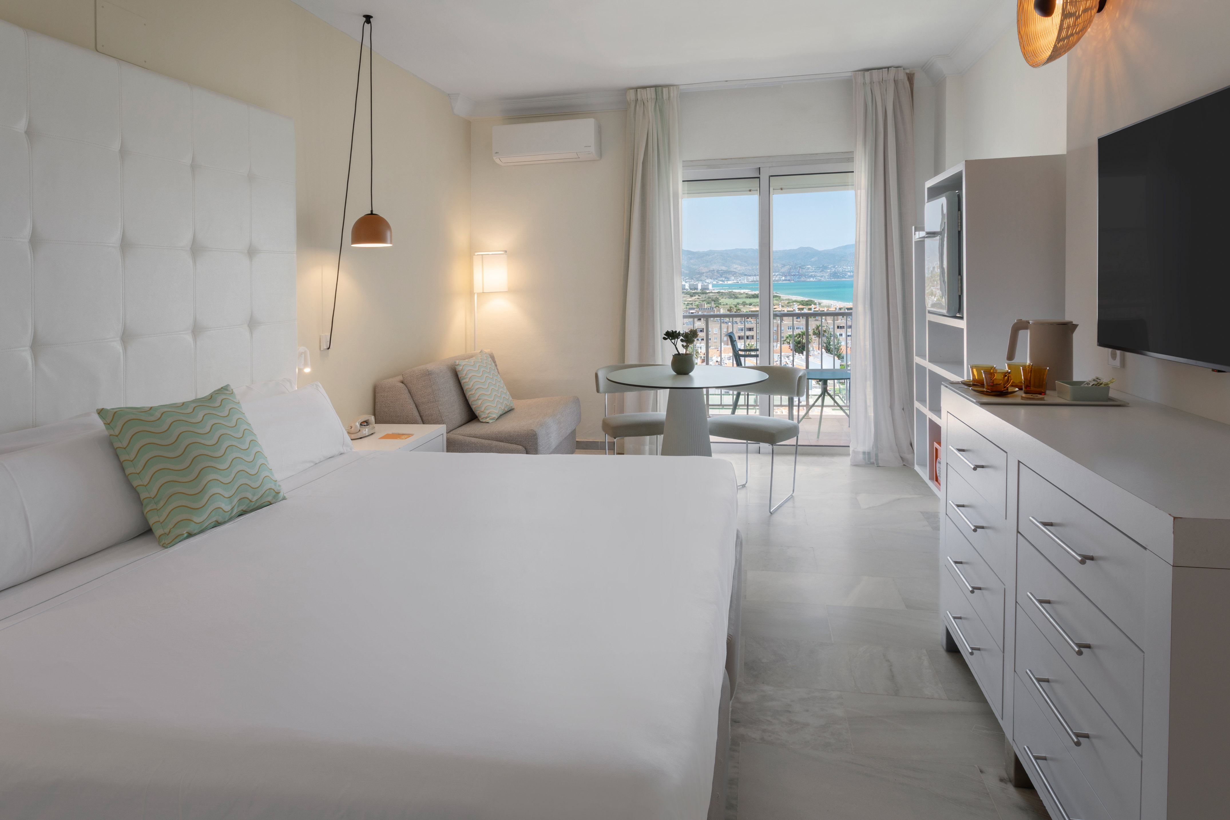 Suite de hotel luminosa con balcón, vistas al mar, cama doble y zona de estar.