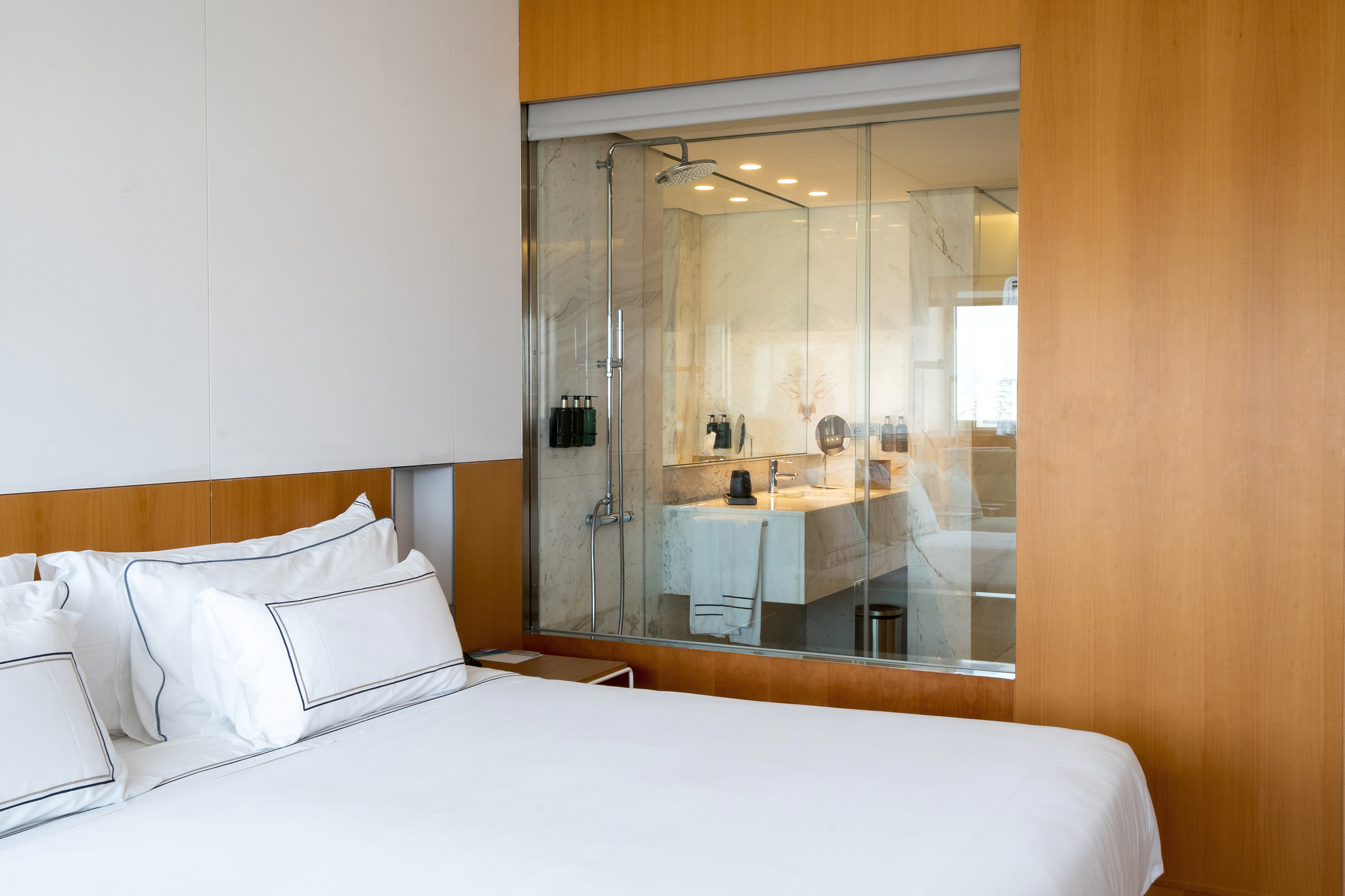 Modern hotel room with white bed, glass-enclosed marble bathroom.