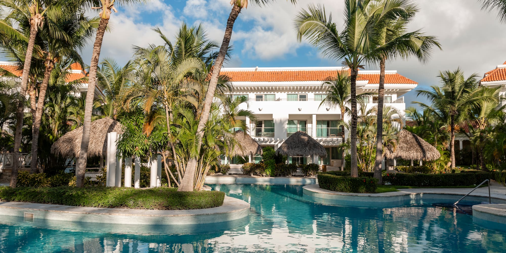 Tropical resort pool framed by palm trees, white hotel, and thatched cabanas.
