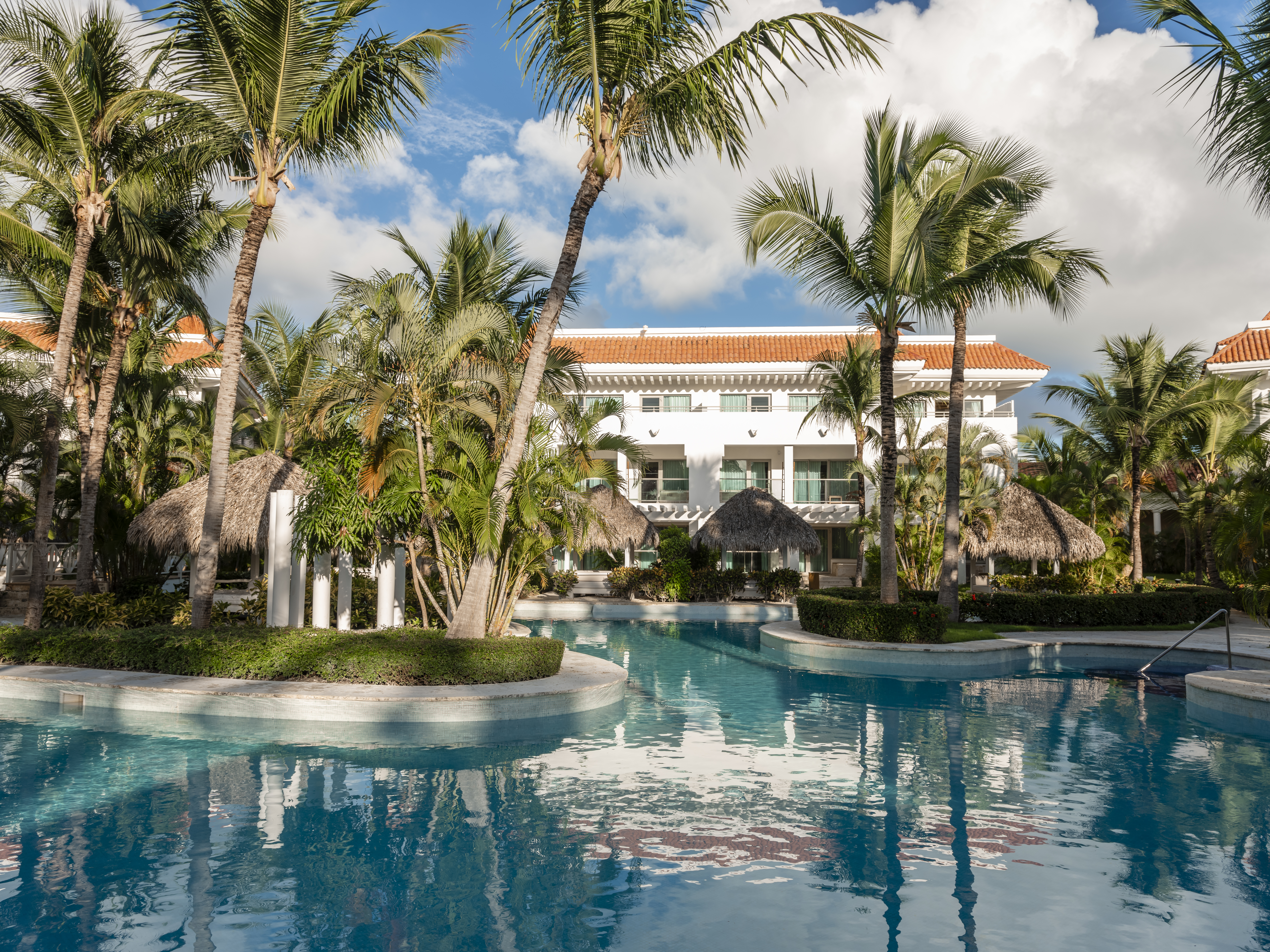 Tropical resort pool framed by palm trees, white hotel, and thatched cabanas.