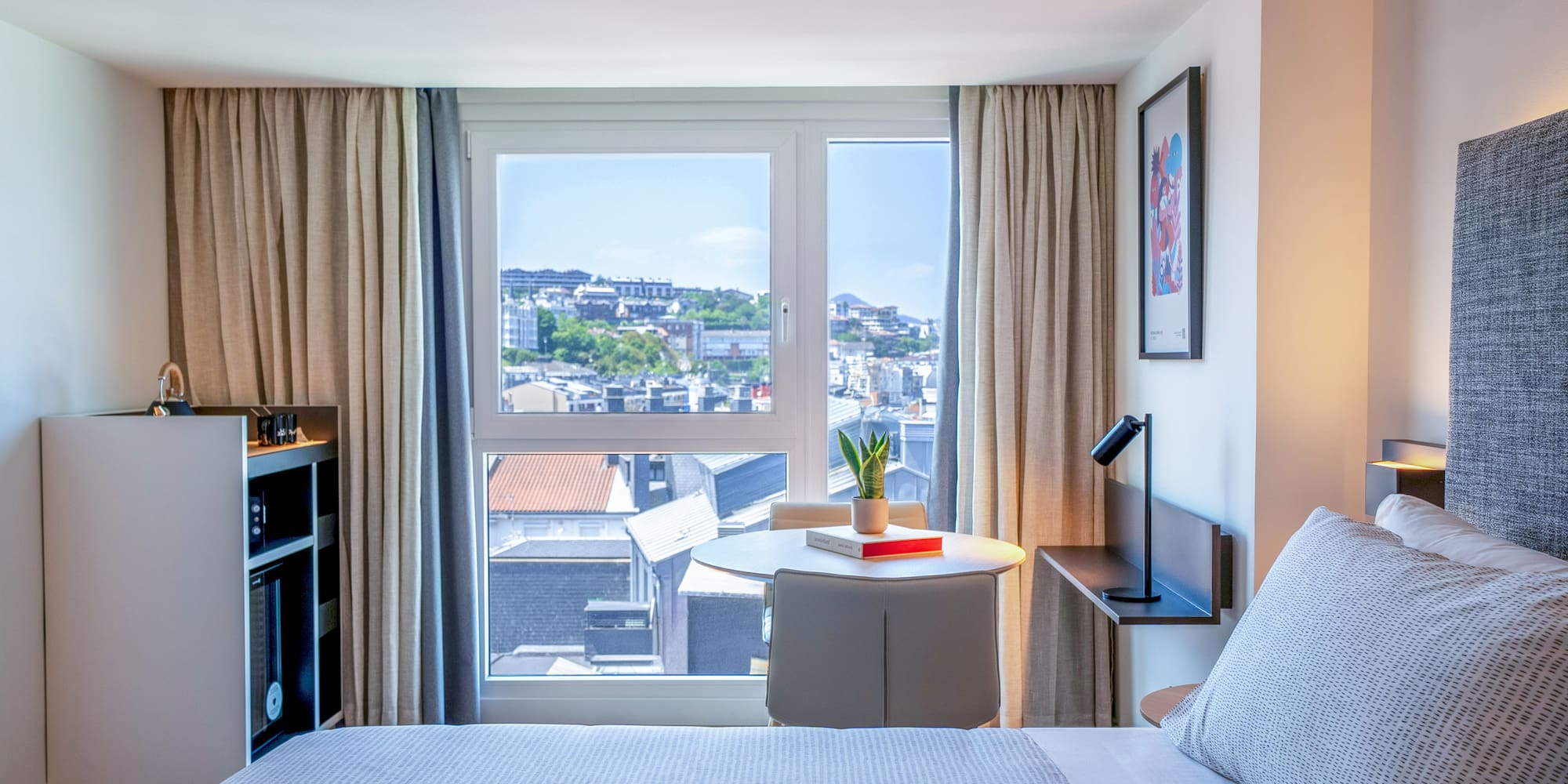 Habitación hotel moderna con vista urbana, cama y mesa junto a la ventana.