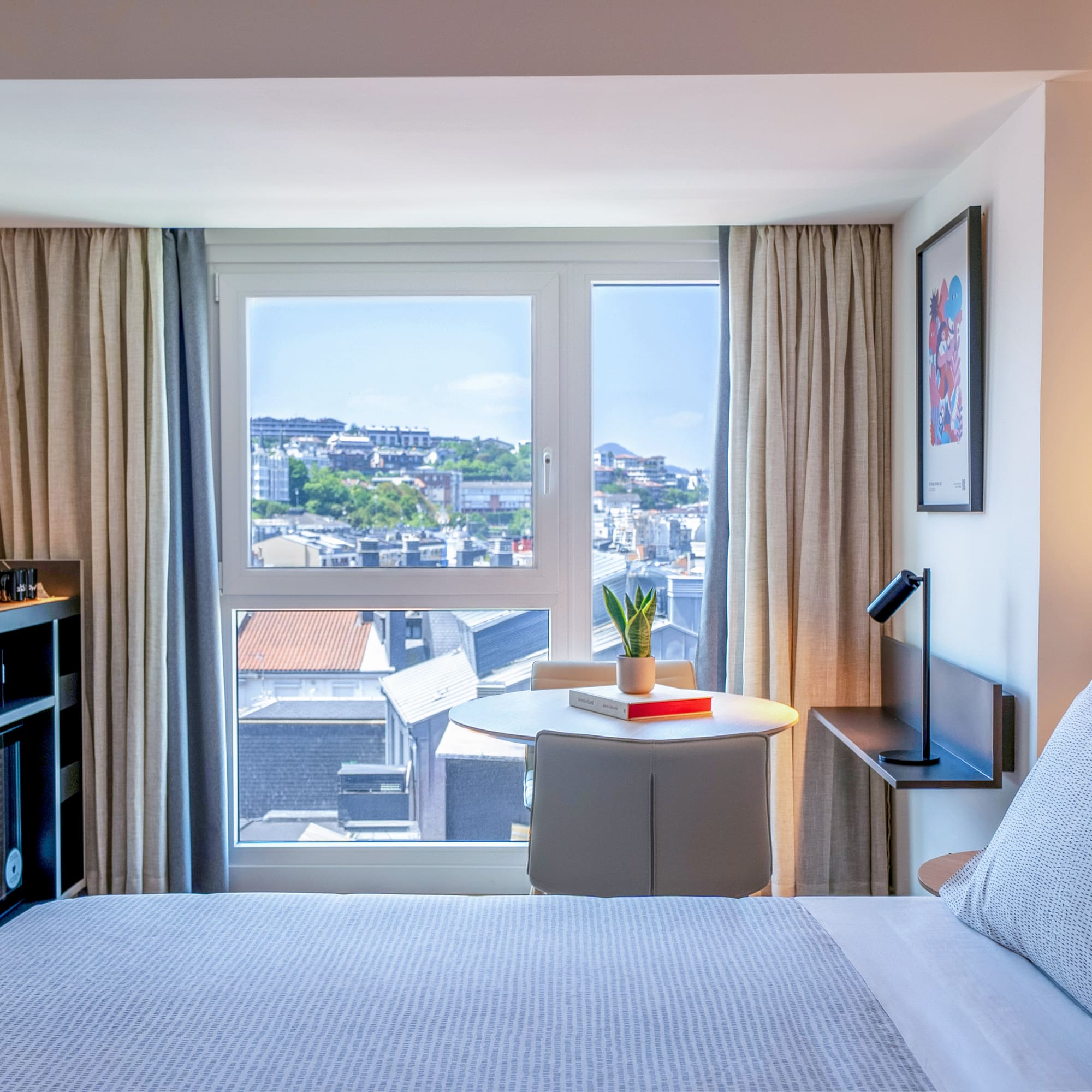 Habitación hotel moderna con vista urbana, cama y mesa junto a la ventana.