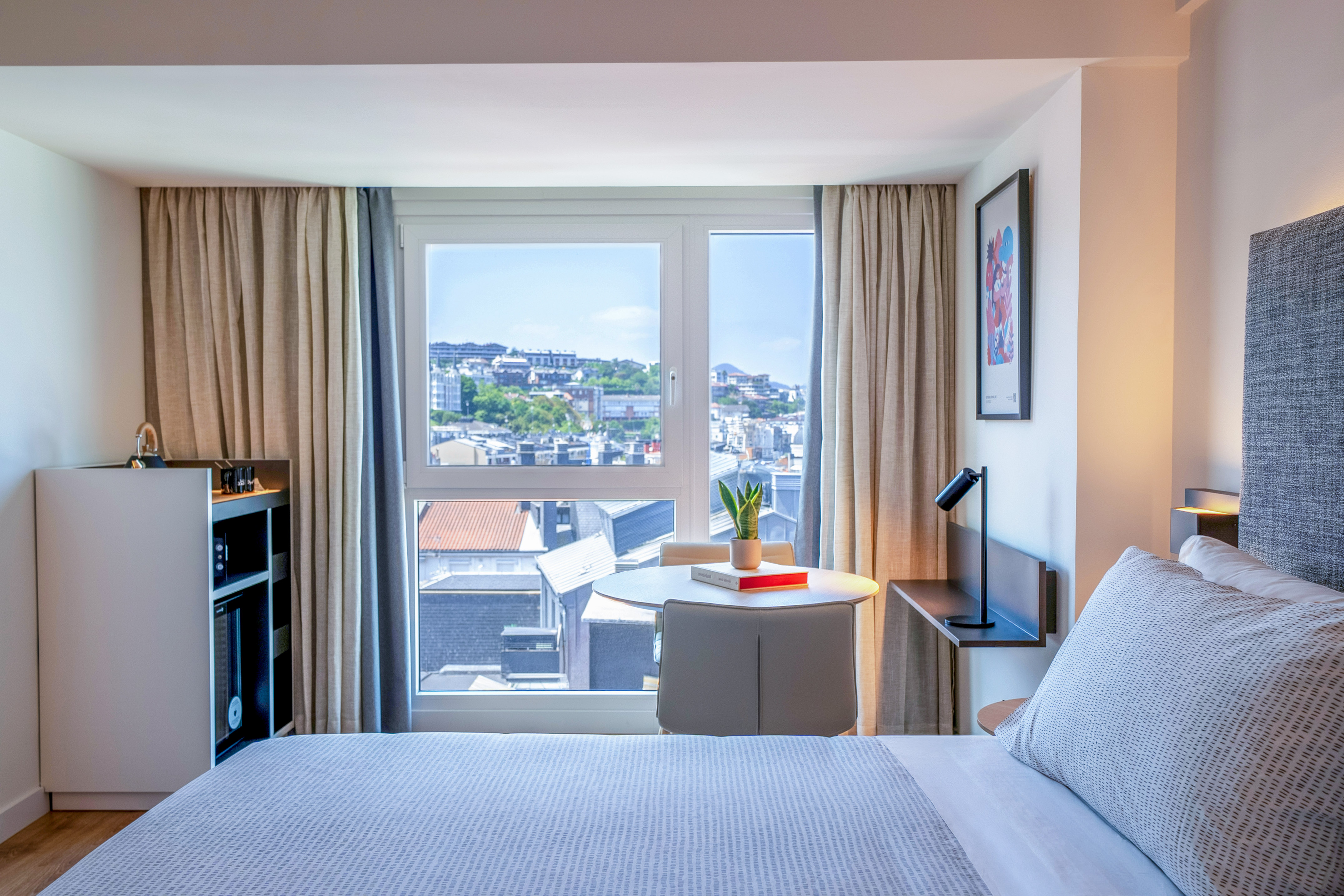 Habitación hotel moderna con vista urbana, cama y mesa junto a la ventana.