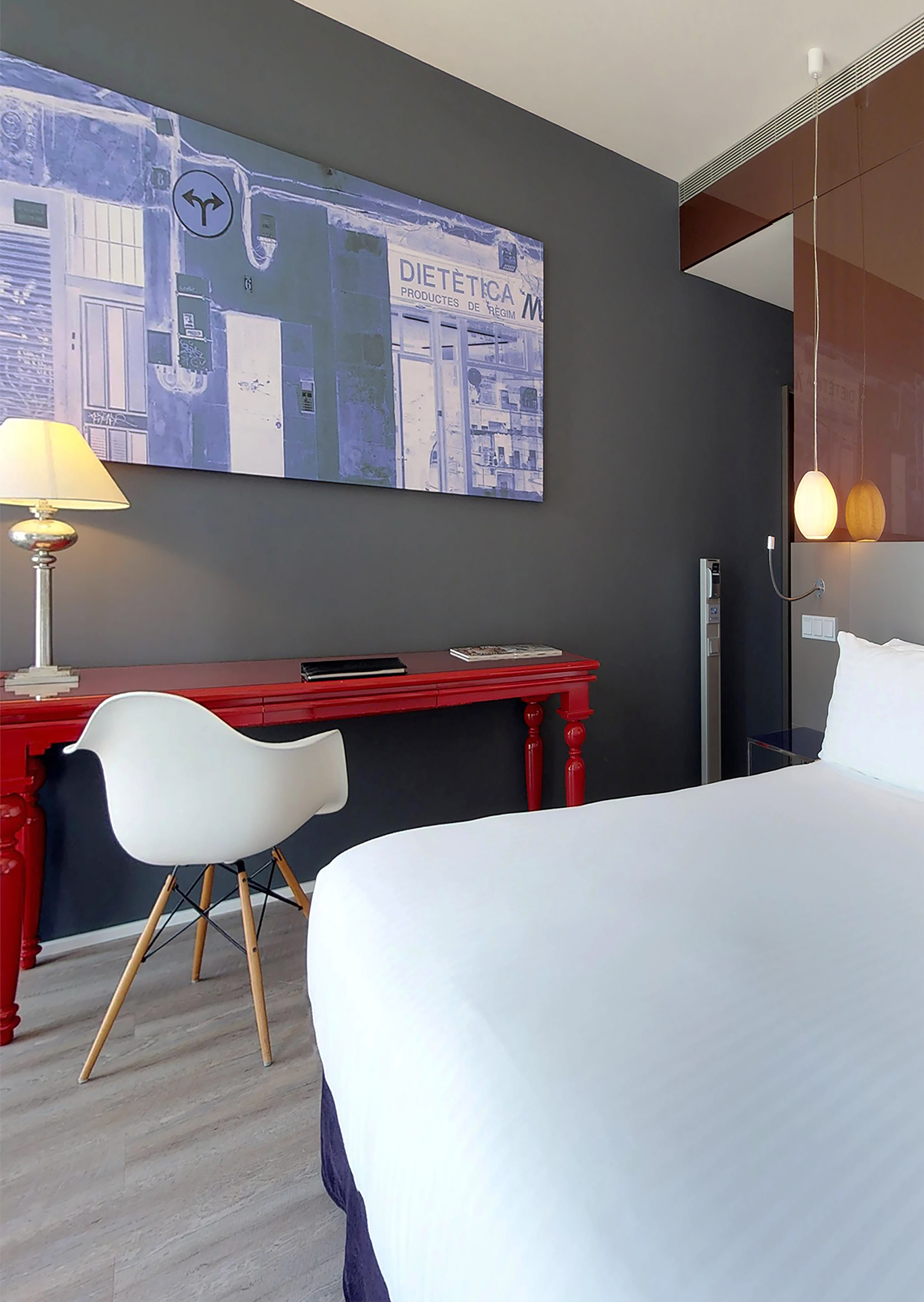Hotel bedroom with white bed, red desk, modern chair, and large urban wall art.