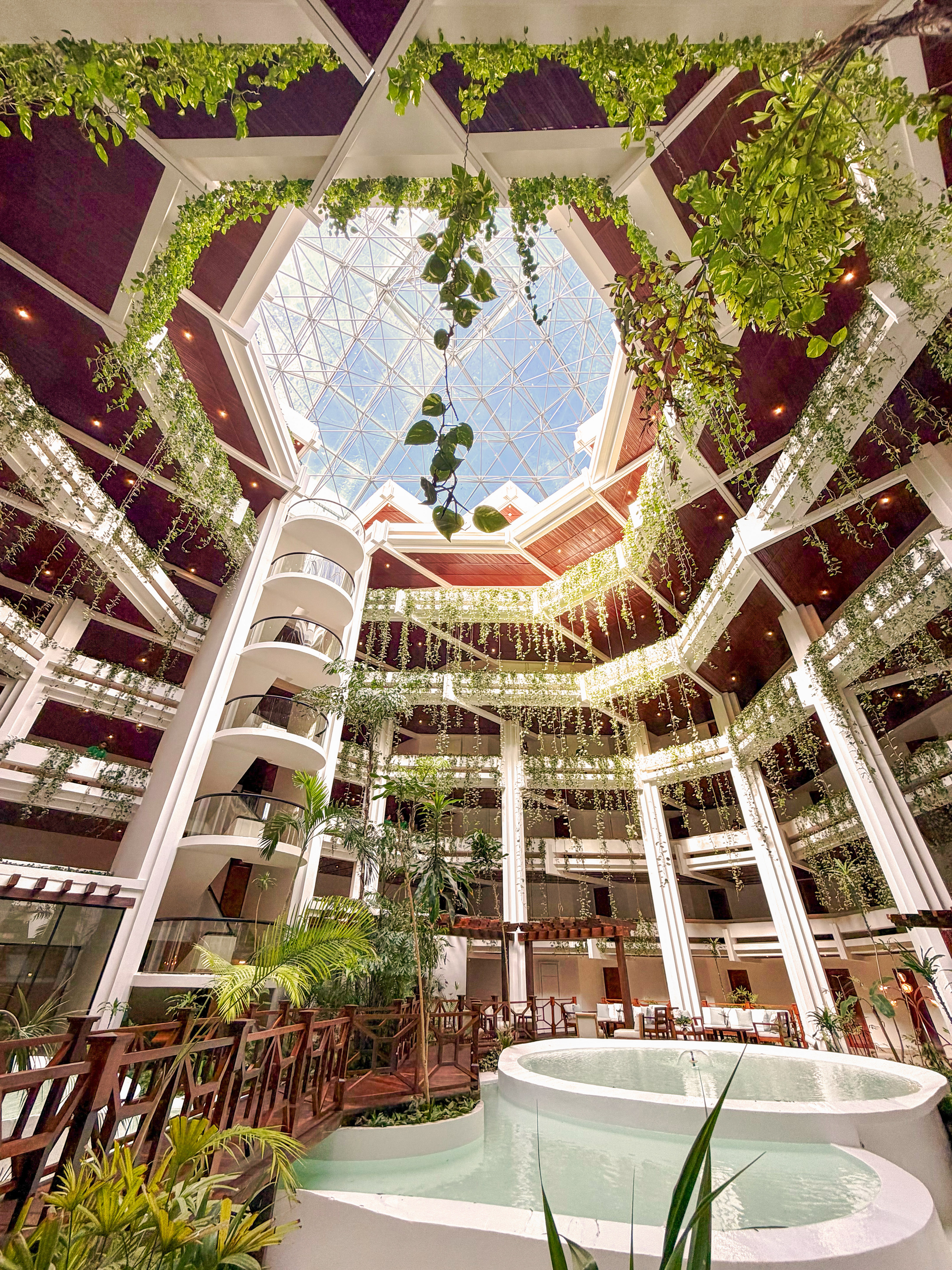 Lush hotel atrium with glass skylight, tropical plants, and indoor water feature.