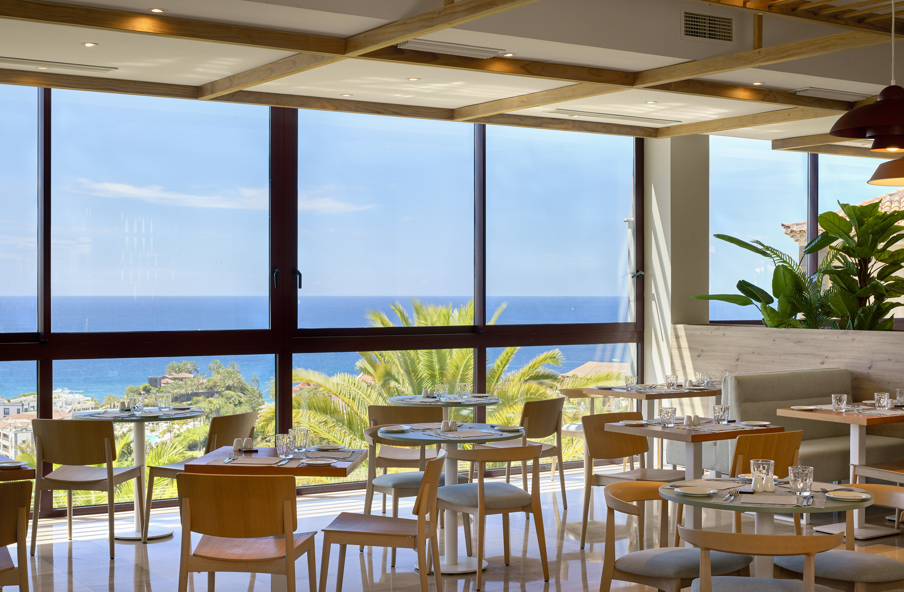 Modern restaurant interior with ocean view, set tables, and palm trees.