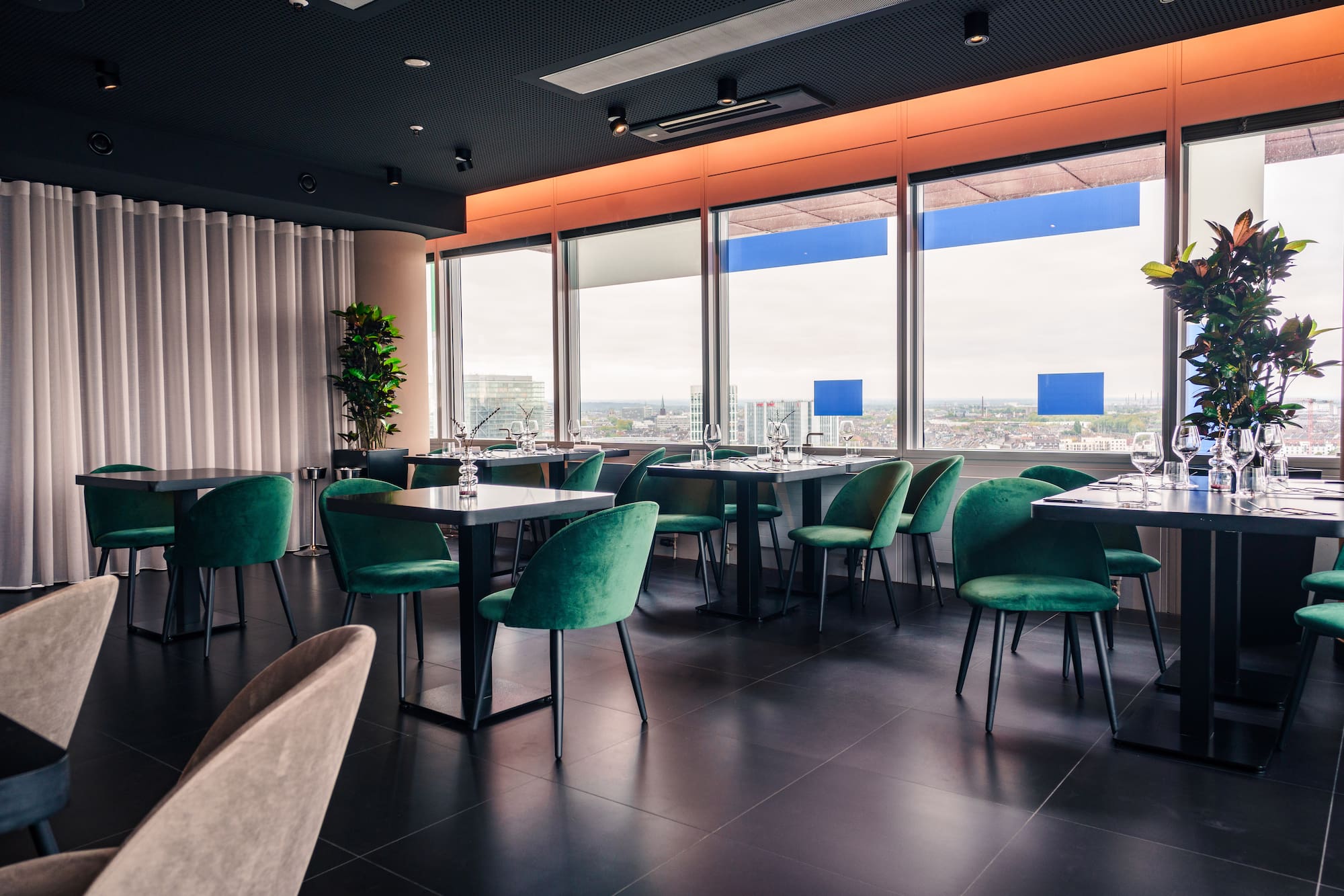 Modern restaurant interior with city view, dark tables, and green velvet chairs.