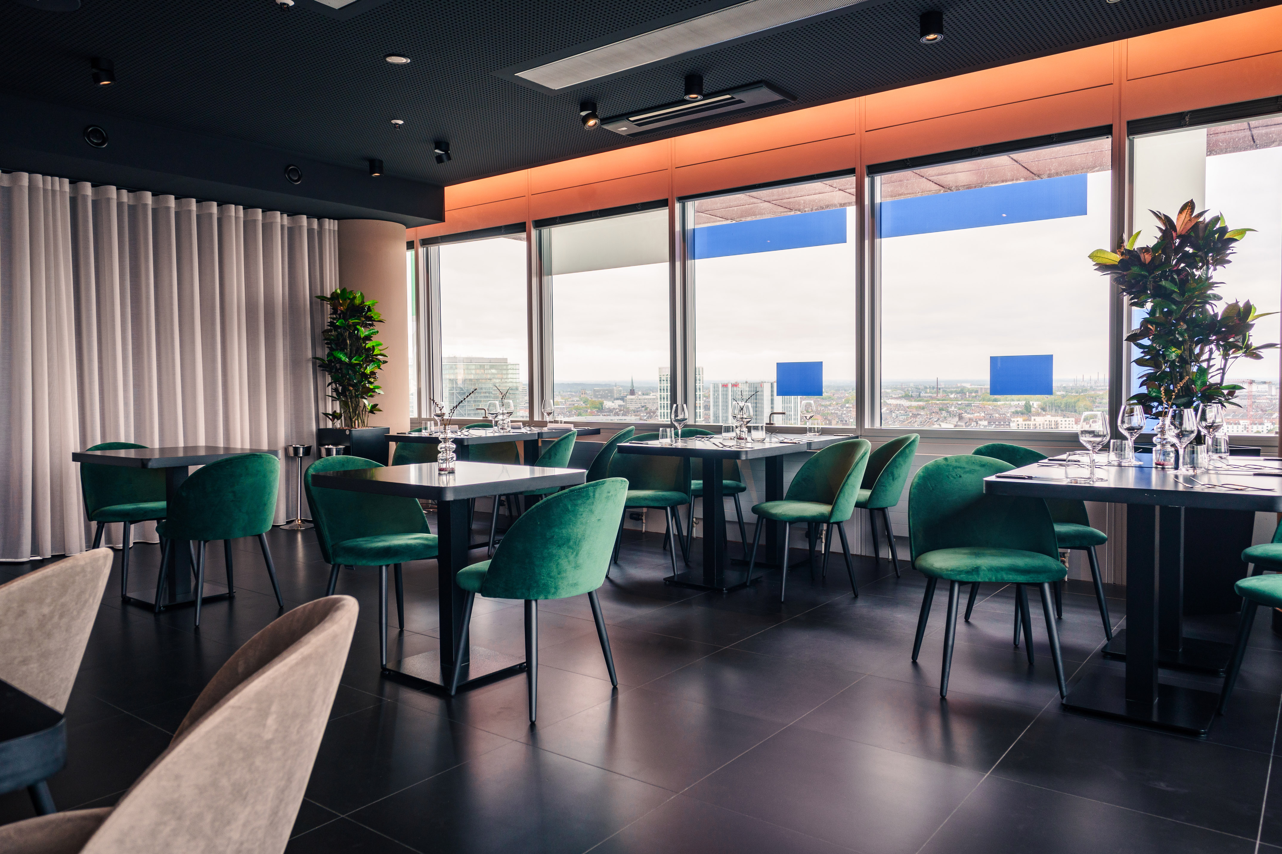 Modern restaurant interior with city view, dark tables, and green velvet chairs.