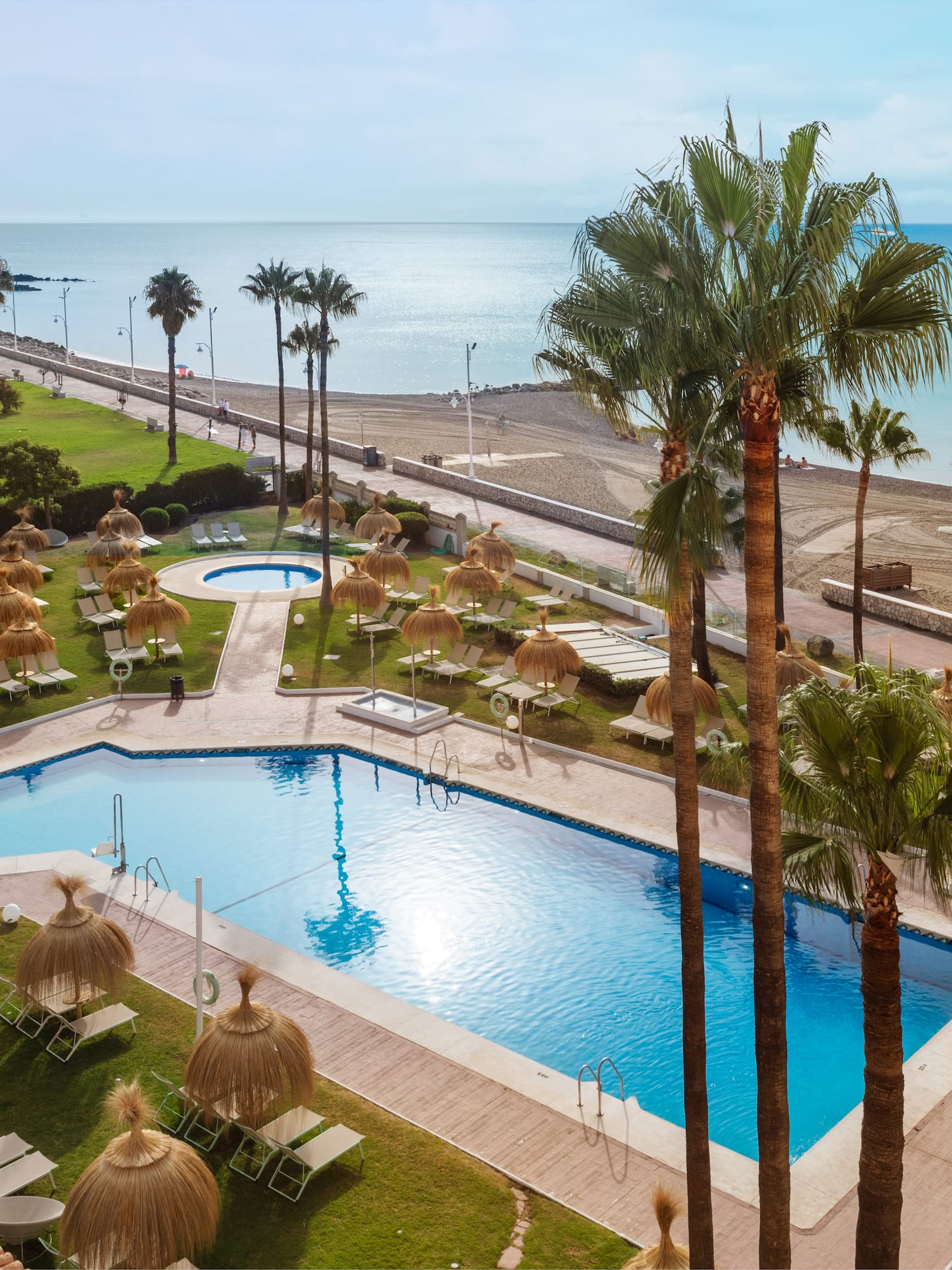 Beach resort pool, palm trees, straw umbrellas, and sandy ocean beach.