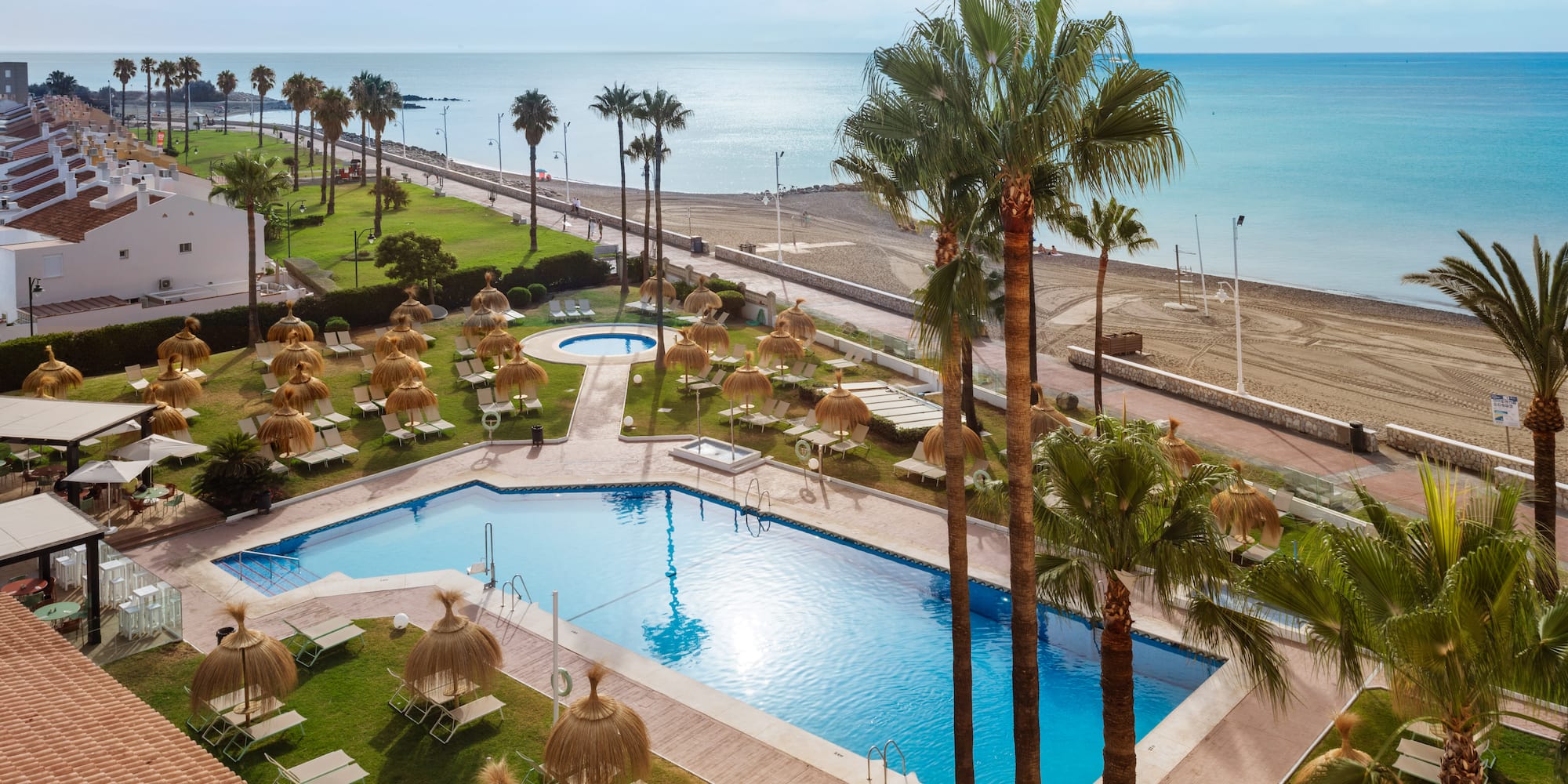 Coastal resort with large pool, palm trees, sun loungers, and sandy beach.